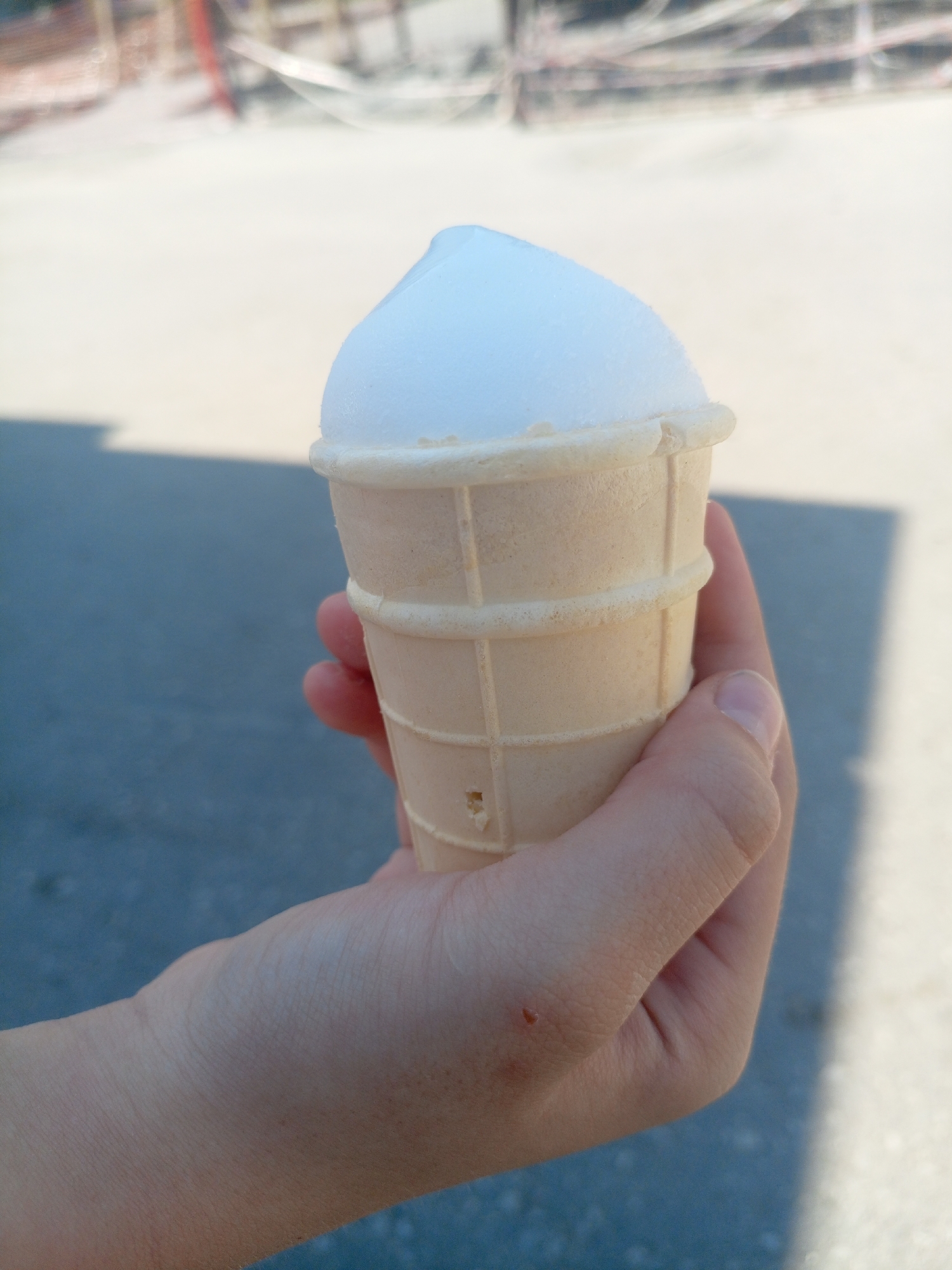 A hand holds a vanilla ice cream cone with white ice cream against a blurred outdoor background of a paved area, possibly a street or sidewalk, with a shadow cast by the ice cream cone and hand on the lighter toned pavement. In the upper portion of the image, a blurry structure with red and white elements, resembling construction scaffolding or fencing, is visible. The lighting suggests it is daytime. The image does not contain any specific landmarks or identifying features that would indicate its location within Saratov, Russia.