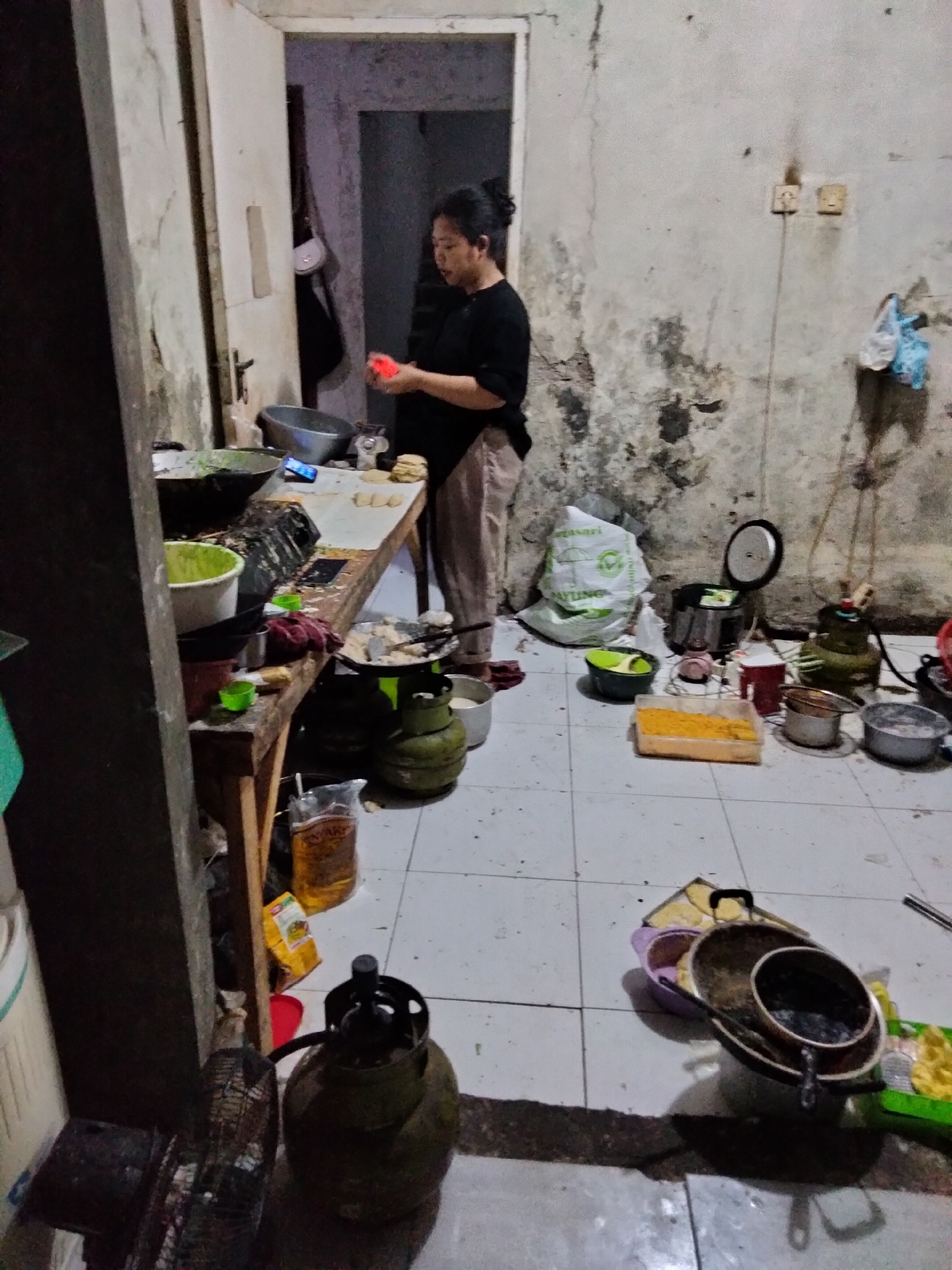 A woman is standing in a dimly lit, cluttered kitchen. The walls are grimy and peeling, and the floor is tiled. A wooden table is laden with cooking utensils and ingredients, including pots, pans, bowls, and a gas cylinder. The woman is wearing a dark shirt and light-colored pants, and she is holding a piece of orange material in her hands. There are several more gas cylinders on the floor, as well as a fan and a rice cooker. The overall impression is one of a humble, working kitchen. The location is identified as Cariu, Indonesia.