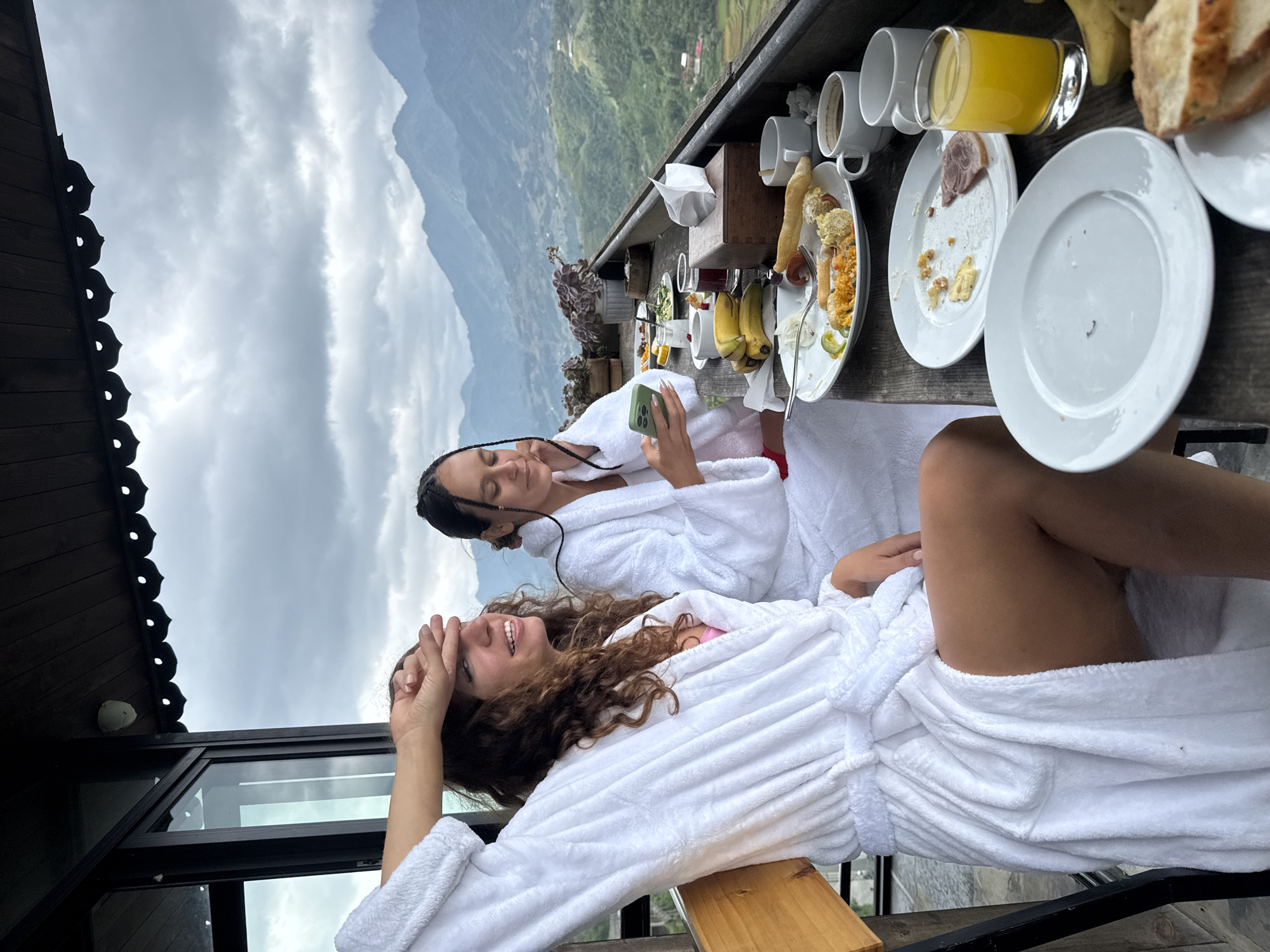 Two women are on a balcony in Sa Pa Ward, Vietnam. They wear white bathrobes. One woman is looking at her phone. The other is smiling, with her hand to her forehead. On the table are leftover plates, cups, glasses of orange juice, bananas, and bread. In the background, there is a cloudy sky and terraced hills.