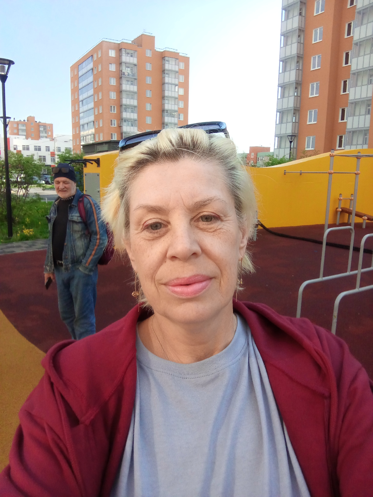 A fair-skinned woman with short blonde hair, wearing a grey t-shirt and a maroon hoodie, takes a selfie outdoors. A man in a denim jacket, black shirt, and black cap stands in the background, holding a mobile phone. The setting appears to be a playground with a yellow wall and metal exercise bars. Behind the playground, there are apartment buildings characteristic of urban residential areas. Based on the architecture and available information, the location is likely in Kommunar, Russia.