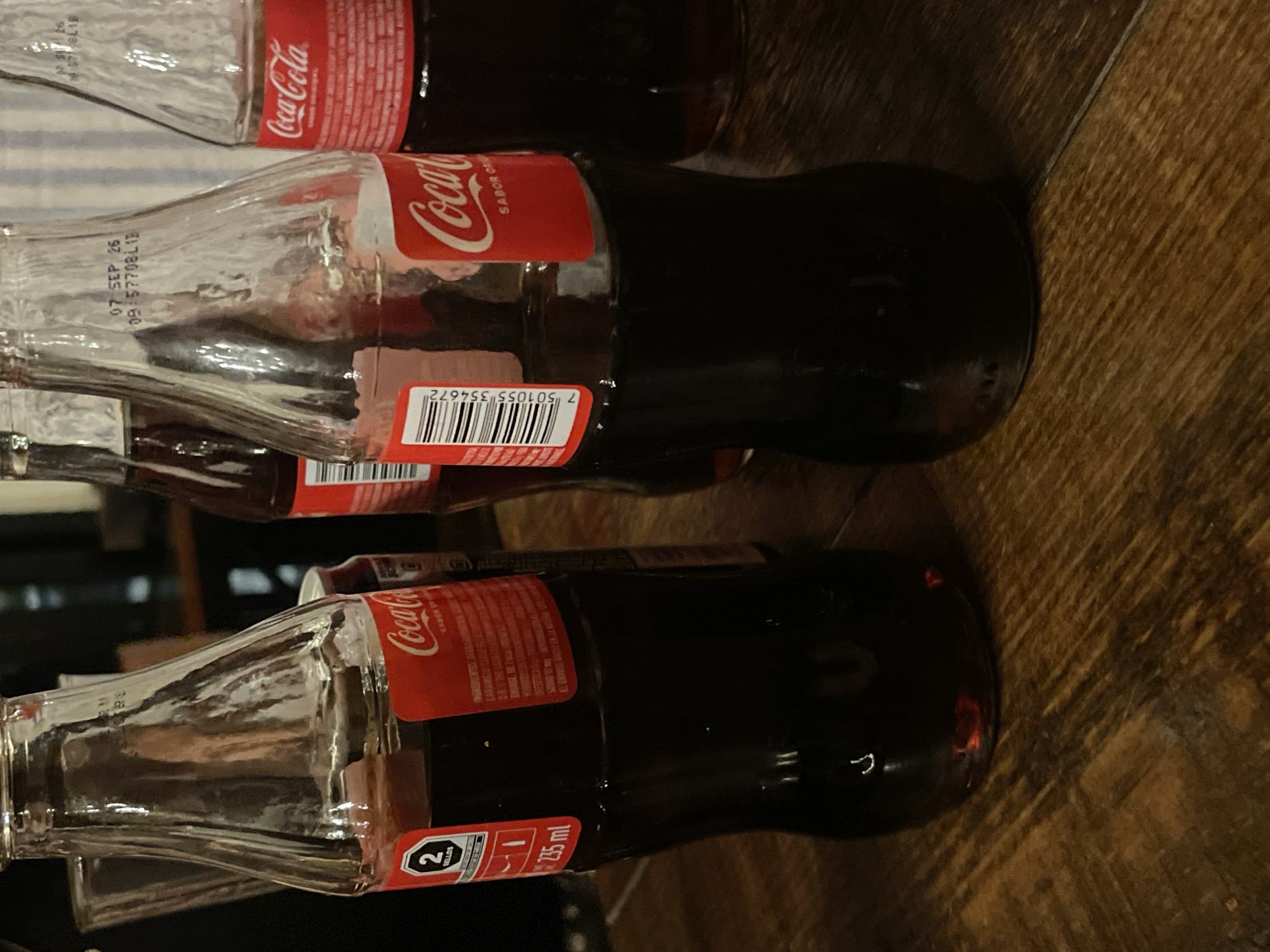 The image captures several full glass Coca-Cola bottles resting on a dark brown wooden surface, possibly a table or floor. The bottles are laid on their sides, with the camera oriented to show them vertically. The lighting is dim, creating reflections on the glass and casting a warm hue on the wooden grain.
The subjects are classic Coca-Cola contour bottles, each containing the dark brown soda. No activity is taking place; the image is a still life.
Notable details include the dark and rich appearance of the wooden surface and the way light interacts with the glass bottles. The labels are predominantly red with white text. The visible text on the bottles confirms they are "Coca-Cola" with "SABOR ORIGINAL" (Original Flavor). The volume "235 ml" is stated, and a black octagonal warning label indicating "2 SELLOS" (2 Seals) is present, a feature common on products in Mexico due to nutritional regulations. A barcode "7 501055 354672" is also visible. Etched onto the glass of one bottle is a date stamp "07 SEP 26" and a batch code "09:57708L1B". Other partial text on labels includes "INGRINEWTIES" (likely "INGREDIENTES" - ingredients), "CARAMELC" (likely "CARAMELO" - caramel), and "ENVASE SE" (likely part of a phrase about the container). The presence of Spanish text and the "SELLOS" labels are consistent with the provided location context of Puebla, Mexico.