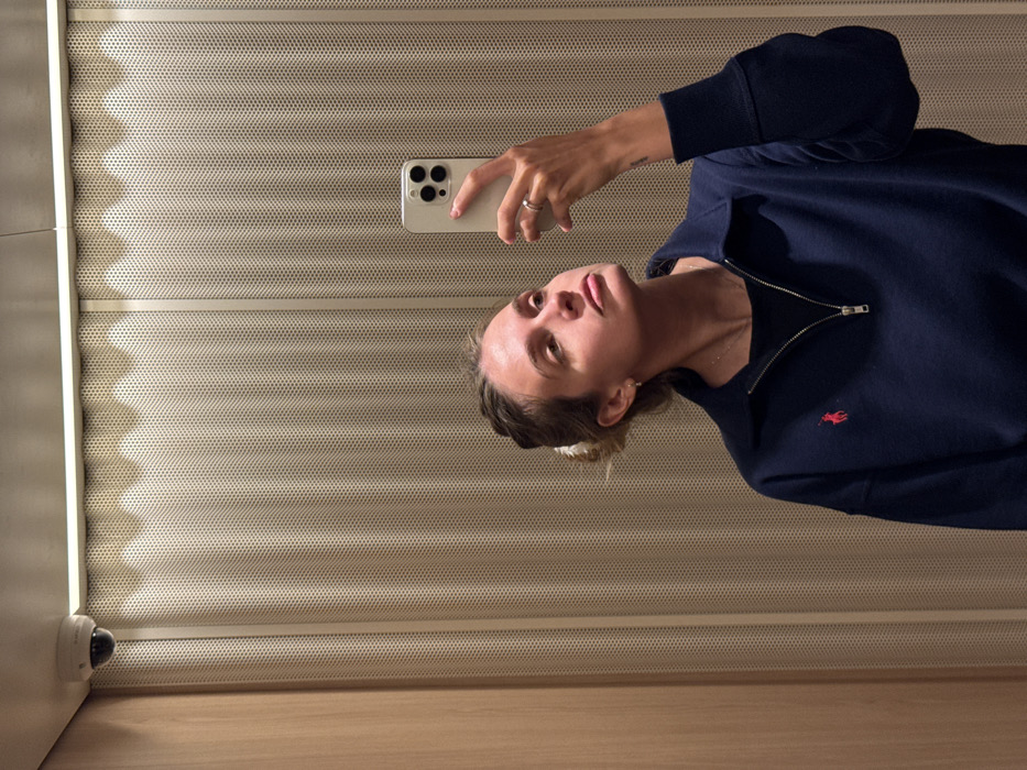 An indoor scene, likely an elevator or a confined modern space, is depicted in a mirror selfie. The location is an unspecified modern building within the city of Tbilisi, Georgia.
A single female subject with light skin and light brown hair, pulled back with a white claw clip, is visible from the chest up. She looks upward and slightly to her left, displaying a neutral expression with a subtle pout. She wears a navy blue quarter-zip sweatshirt featuring a red Polo Ralph Lauren pony logo on the left chest, layered over a black undershirt. A thin silver necklace and a silver ring on her left ring finger are visible. A small tattoo is discernible on her right forearm/wrist.
Her right hand is raised, holding a light-colored smartphone (consistent with an iPhone model featuring three rear camera lenses) vertically, pointed towards herself to capture the reflection.
The background consists primarily of light beige or tan perforated metal wall panels, creating a textured pattern. A section of smooth, light-colored wooden paneling is visible on the far left. A linear LED light strip runs horizontally along the upper wall/ceiling junction, illuminating the space. A white dome-shaped security camera is mounted in the upper left corner of the frame, near the ceiling. The subject is actively performing the action of taking a selfie.