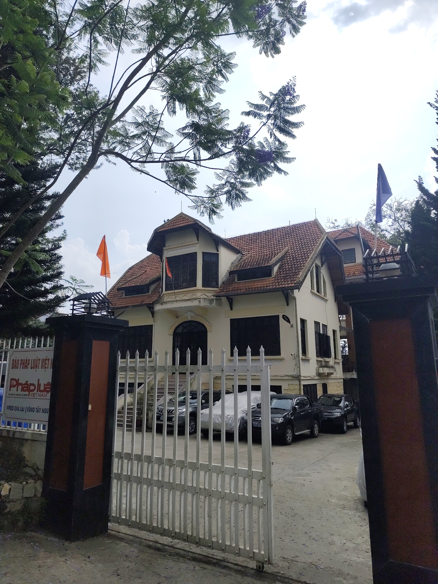 The image shows a three-story building with a terracotta-tiled roof, located in Da Lat, Vietnam. The building has a white facade with dark window shutters and a central balcony. A white metal gate is in the foreground, partially obscuring several vehicles parked within the building's courtyard. A sign with Vietnamese text, including the words "Pháp luật Việt Nam," is visible on the left gatepost. An orange flag is visible to the left of the building, and a blue flag is seen on the right. Trees partially frame the image, with some branches obscuring the sky above.