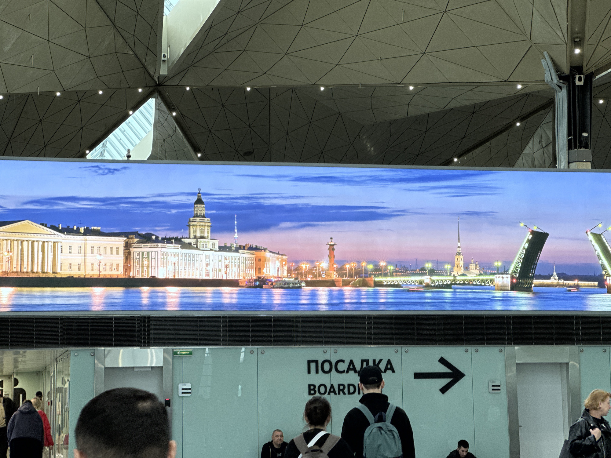 The image depicts an airport interior, likely in Saint Petersburg, Russia, evidenced by the large LED screen showcasing a panoramic view of the city, featuring landmarks such as the Palace Embankment, the Admiralty Building, the Rostral Columns, and the Palace Bridge raised at night. Below the screen, boarding signage in both Russian ("ПОСАДКА") and English ("BOARDING") is visible, accompanied by a right-pointing arrow. Several people are present, including individuals walking towards the boarding area and others seated or standing near the wall. The architecture overhead consists of a complex geometric ceiling structure with integrated lighting.