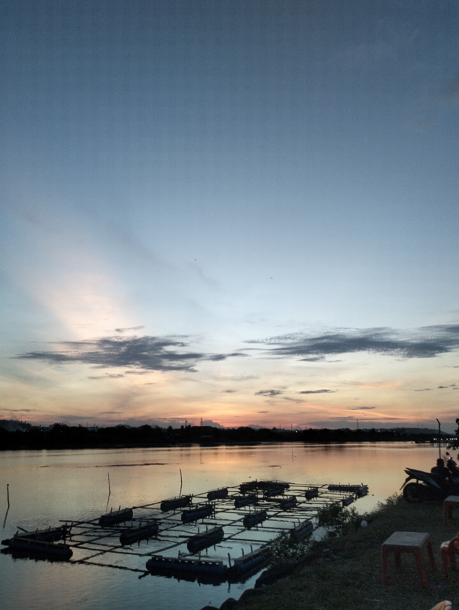 The image depicts a sunset over a body of water, likely a lake or river, in Lhokseumawe, Indonesia. The sky is filled with varying shades of blue, gray, and orange, indicating a setting sun. A dark silhouette of land is visible in the distance. In the foreground, a collection of floating rafts or platforms are arranged in a grid-like pattern on the water's surface. On the right side of the frame, the edge of the land is visible, with grass and a few pieces of furniture, including a motorcycle and stools, situated on the shoreline. The overall scene appears peaceful, capturing the stillness of the water and the fading light of dusk.
