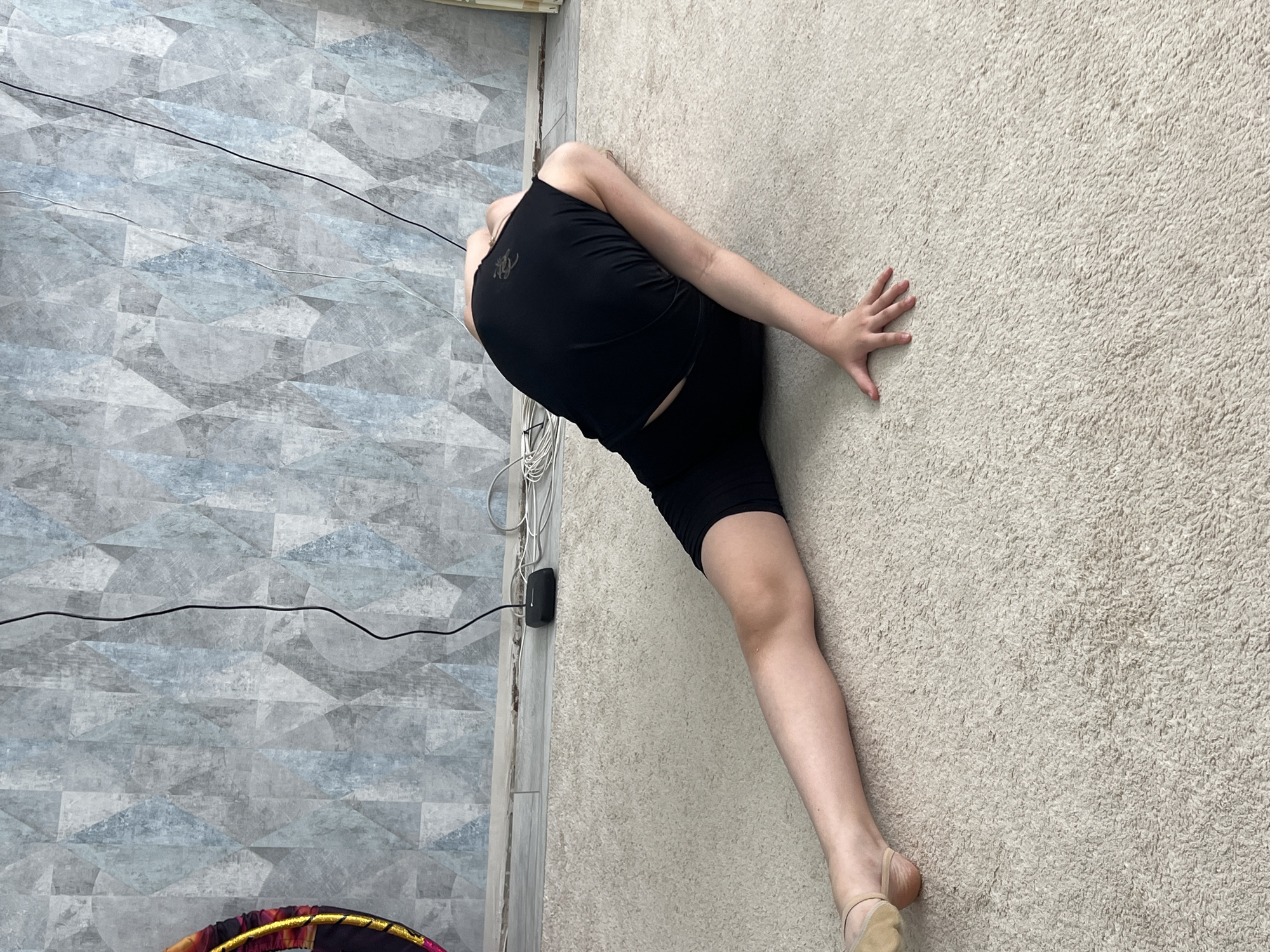A gymnast in a black tank top and black shorts is positioned on a beige textured carpet in a room in the city of Chaikovsky, Russia. She is performing a contortion pose, with her right hand pressed against the wall for balance. Her right leg is extended straight out, while her body is bent backwards. Her left leg is bent back behind her. Her head is obscured. A black wire and several white wires are running along the wall on the left side of the image, near a power outlet. The wall to the left of the wires has a gray geometric pattern. A portion of a multi-colored hula hoop is visible in the bottom left corner.