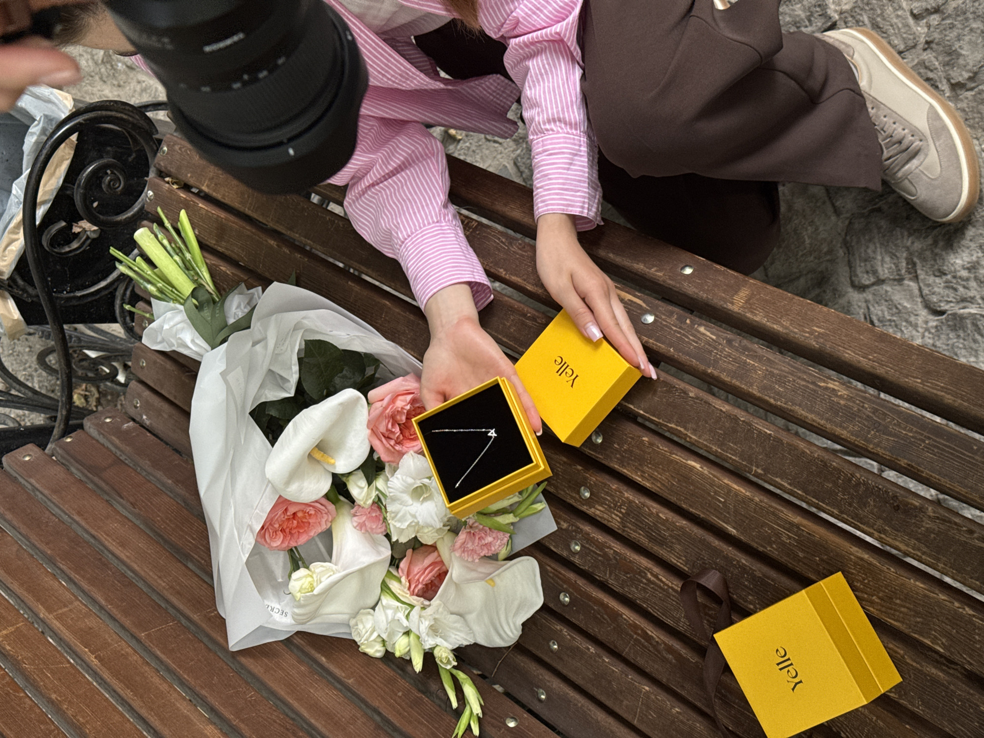An overhead shot captures a person either setting up or capturing a moment on a wooden slatted bench outdoors. The person, dressed in a pink and white striped shirt, brown pants, and grey sneakers, is partially visible. Their left hand is seen holding a black camera lens, indicating a photography activity.
On the bench, a beautifully arranged bouquet of flowers wrapped in white paper takes center stage. The bouquet features prominent white calla lilies with yellow pistils, delicate pink roses, and other smaller white flowers and green foliage. The white wrapping paper has partially visible text, "SECRE".
The person's right hand is positioned over a small, bright yellow box that is open, revealing a slender silver necklace with a small geometric pendant resting on a black insert. Another identical yellow box, closed and adorned with a brown ribbon, is placed further down the bench. Both yellow boxes clearly display the brand name "Yelle" printed vertically on their sides.
The bench is made of dark brown wooden slats, and the ground below is a textured surface of grey stones or cobblestones. The scene suggests a special occasion or a meticulously arranged flat lay for product photography, showcasing gifts of flowers and jewelry. The time of day appears to be daylight, with soft, even lighting.