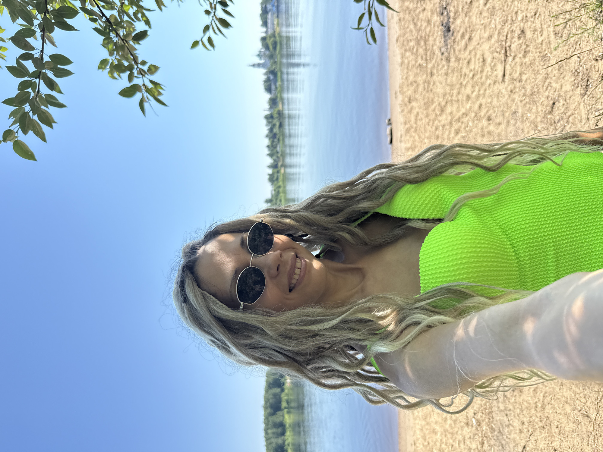 A woman with blonde dreadlocks and sunglasses smiles at the camera, holding it out to take a selfie. She is wearing a neon green swimsuit and stands on a sandy beach with a body of water behind her. In the distance, across the water, is a city with buildings and a church steeple. Trees line the far bank of the water. The sky is clear and blue. The image appears to have been taken in Заволжье, Russia, given the presence of the church steeple and the overall landscape.