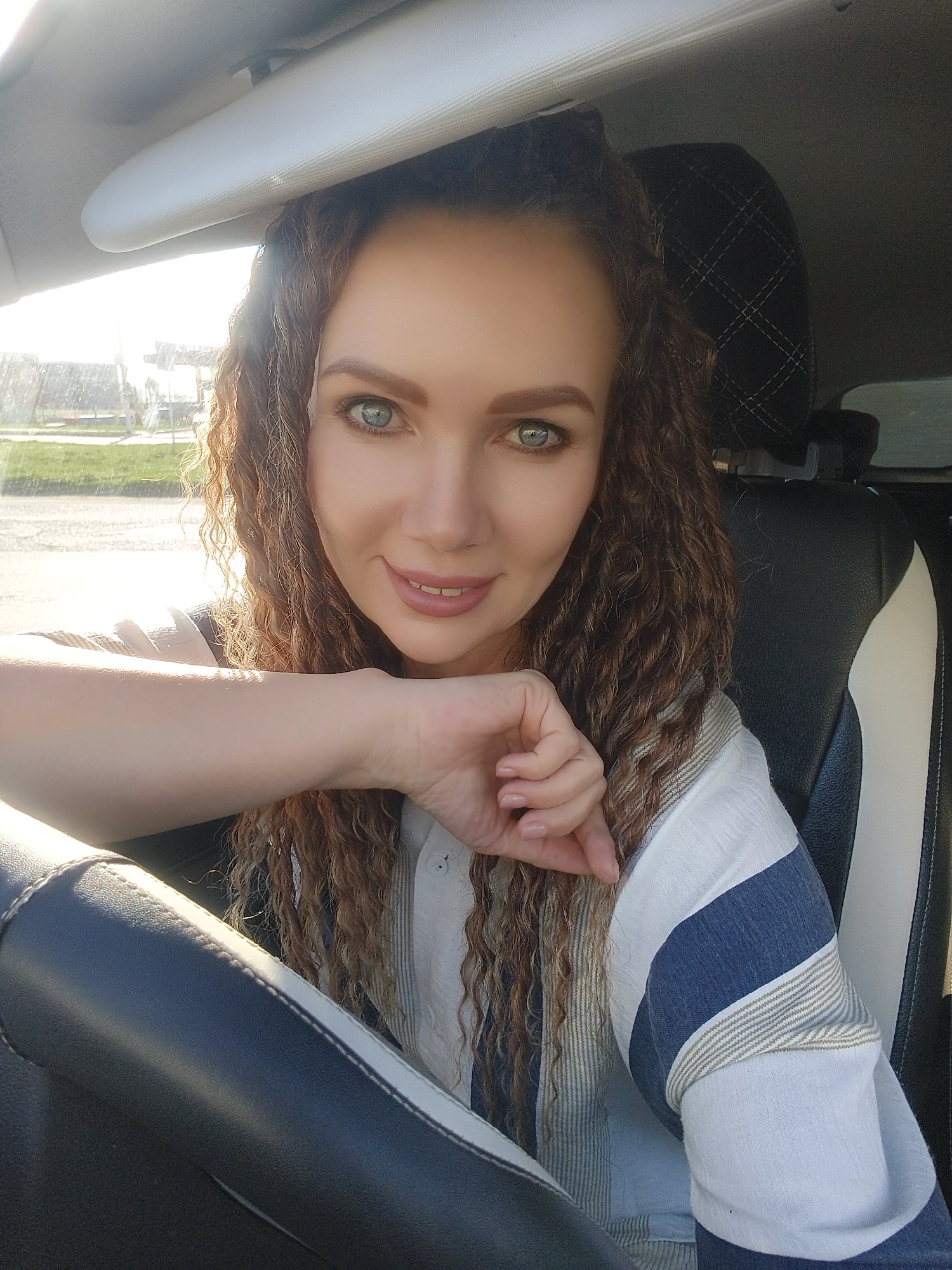 A woman is seated in a vehicle, visible from the chest up. She has long, curly brown hair, fair skin, and light-colored eyes. She is wearing a striped, light-colored shirt with blue stripes. Her left arm is resting on the steering wheel. The steering wheel is dark, likely black, with a light-colored stripe. A car seat, black with a white stripe, is visible behind her right shoulder. Above her head is the interior roof of the car, which is light-colored. The setting appears to be outdoors, with greenery and buildings visible blurred through the window in the background. The location is Unknown, Unknown.