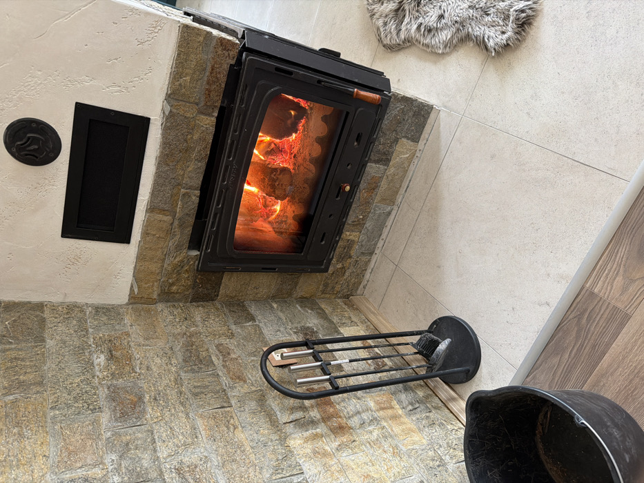 An indoor scene in Panagyurski kolonii, Bulgaria, captures a room corner with a functional fireplace. The fireplace structure is faced with rectangular, multi-toned stone tiles on its lower section and an adjacent left wall, transitioning to textured off-white plaster on its upper section. A black metal fireplace insert with a glass door is centrally positioned, revealing burning logs and active flames within. Above the insert, within the plaster, a black rectangular vent cover and a circular black decorative emblem with a flame motif are visible. The floor in front of the fireplace consists of light-colored square tiles, extending to the right where a light-colored, shaggy rug is partially visible. To the left, the tiled floor meets wood-patterned flooring, separated by a transition strip. A black metal fireplace toolset, comprising a stand holding tongs, a poker, a brush, and a shovel, stands on the tiled floor. A black plastic bucket containing minor debris is partially visible on the wood-patterned floor in the lower left foreground. No individuals are present.