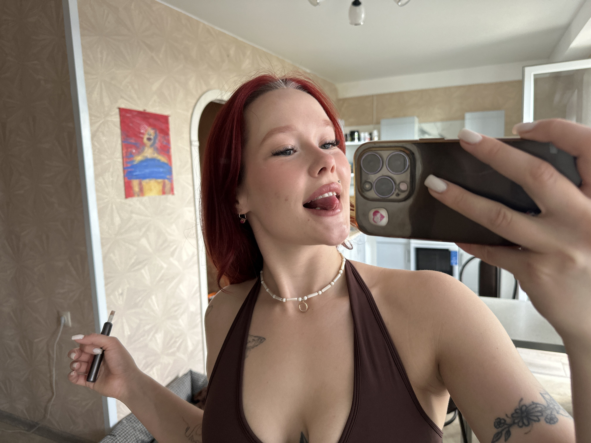 An indoor, eye-level, medium shot shows a young Caucasian woman with red hair taking a selfie with a smartphone. She is wearing a brown halter top and a beaded necklace. Her left arm is extended, holding a gray smoking device. She has tattoos on her left arm and chest. In the background, a framed painting hangs on a textured wall. The lighting is natural, with some artificial light visible from overhead fixtures. The setting appears to be a residential room.