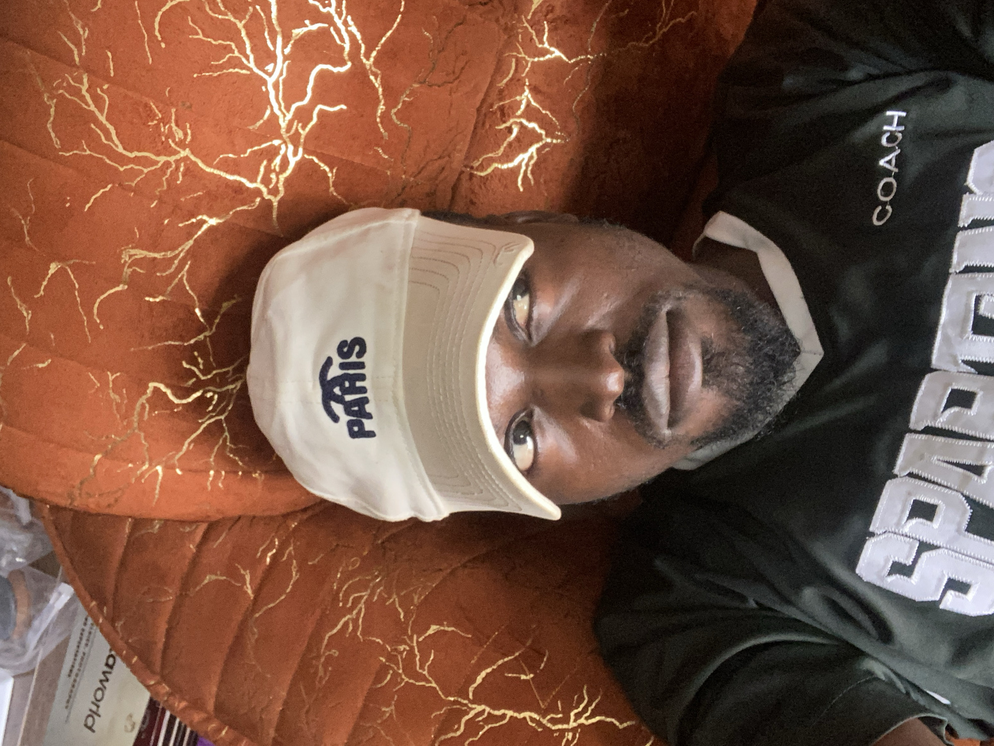 A man wearing a white baseball cap with the word "PARIS" embroidered in blue and a dark green t-shirt with "SPARTAN" and "COACH" written on it, is lying down on an orange patterned surface. The man is looking up towards the camera with his eyes wide open. The background is an orange textured surface with gold-colored, tree-like patterns. There are also some objects visible in the upper left corner, including what appears to be a book or magazine with text in a foreign language. The overall scene appears to be indoors, possibly on a couch or bed. The lighting is dim, suggesting it might be evening or the room is not well-lit. There is no indication of the specific location being Gura Top, Nigeria.
