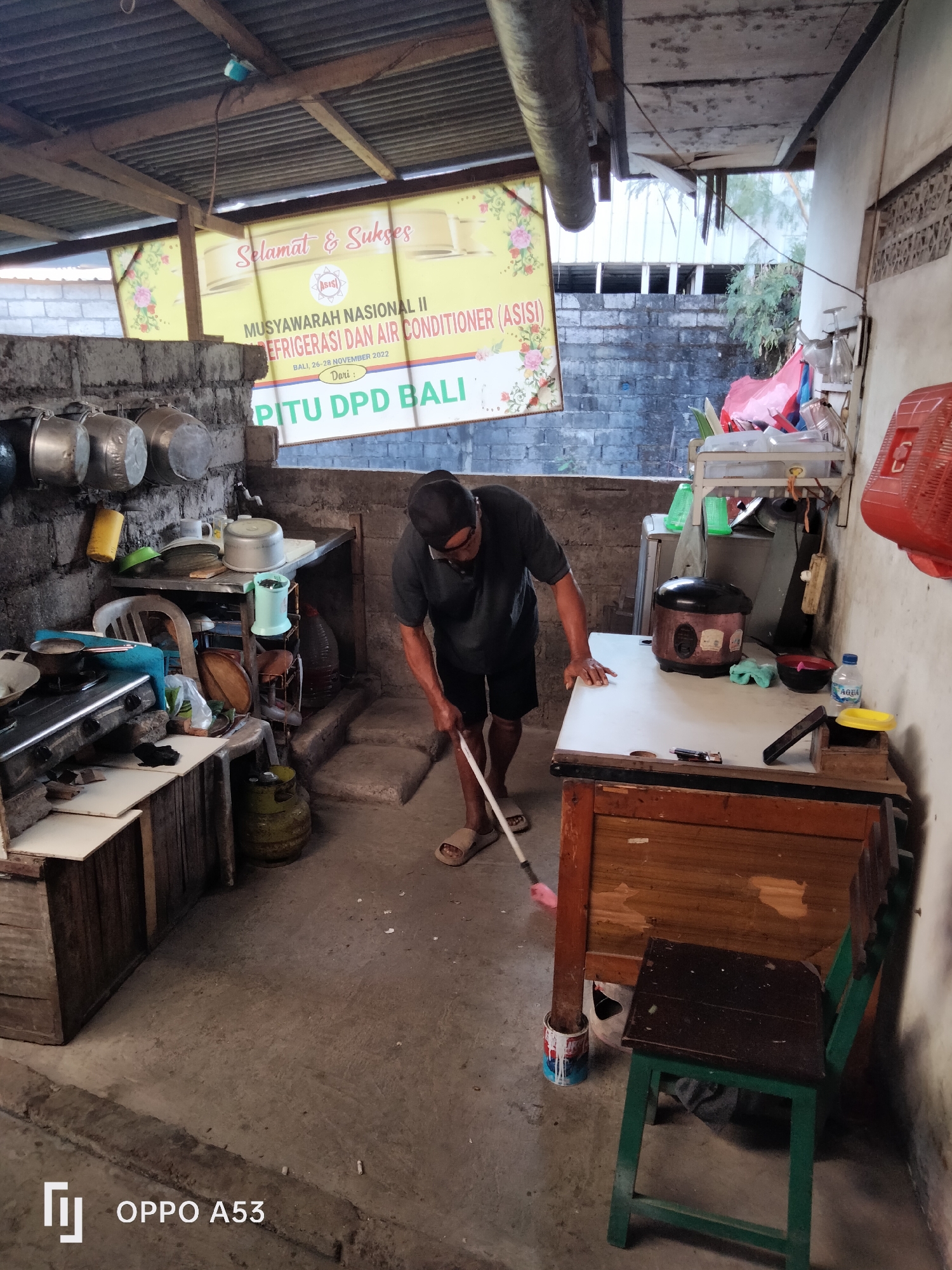 The image depicts an outdoor covered area, possibly a kitchen or workshop, in Denpasar, Indonesia. A man is sweeping the concrete floor with a broom. The environment is cluttered with various objects, including cooking pots hanging on the wall, a gas cylinder, a rice cooker, shelves stocked with items, and a wooden cabinet. A yellow banner with Indonesian text, possibly an advertisement or announcement, is visible on the wall. The overall impression is one of a utilitarian and somewhat unkempt space.