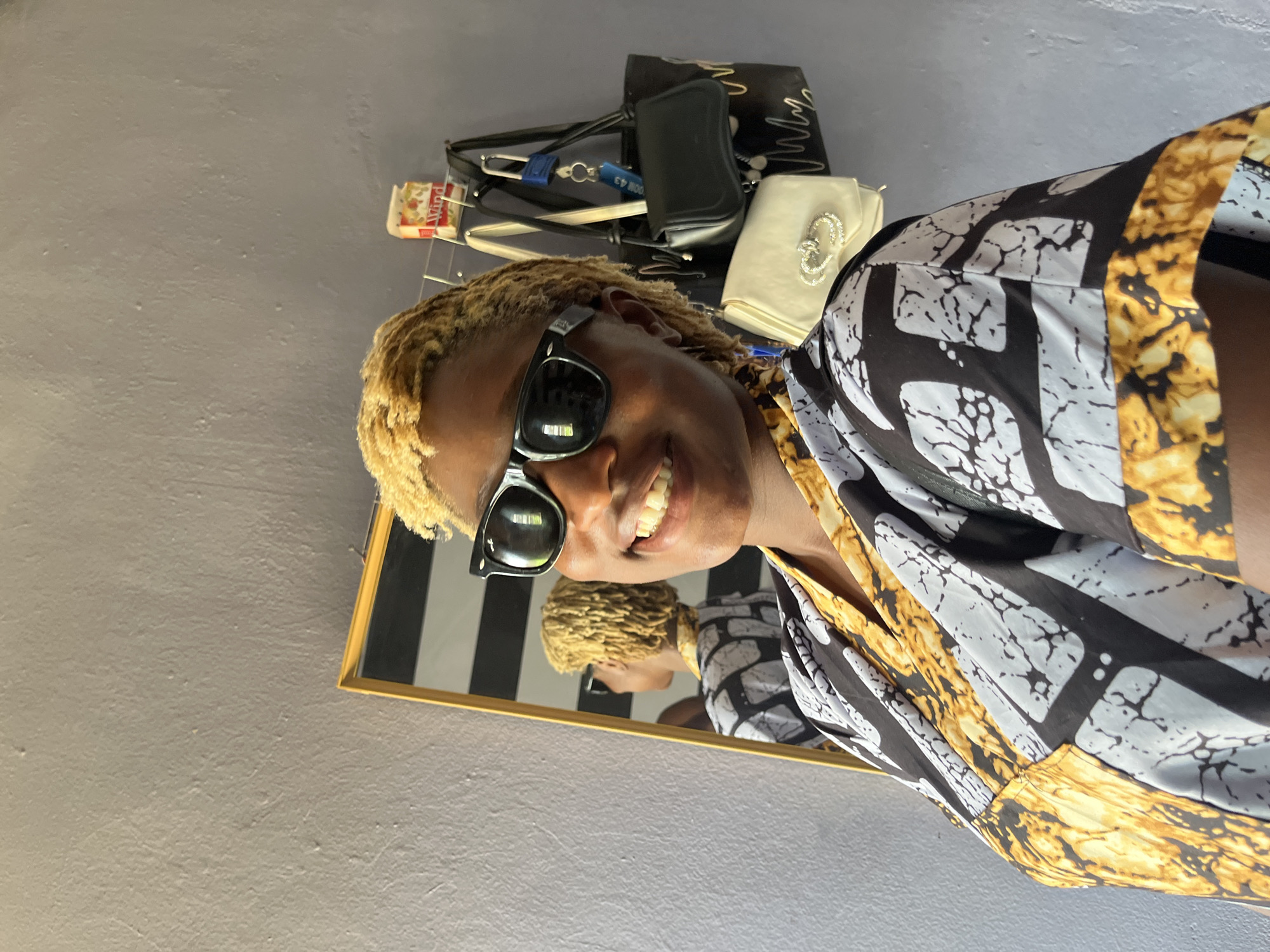 In a medium shot, a young Black woman with short, blonde, dreadlocked hair and sunglasses smiles at the camera, her head tilted slightly to her left. She is wearing a black and white patterned top with gold accents. She is standing in what appears to be a room in Jos, Nigeria, with a gray, textured wall behind her. To her right, a mirror reflects her image, showing her from the back. Several handbags are hanging on a rack behind her, including a black shoulder bag and a small white clutch with a jeweled clasp. A box labeled "Wind" is also visible on the rack. The lighting is bright, casting soft shadows.