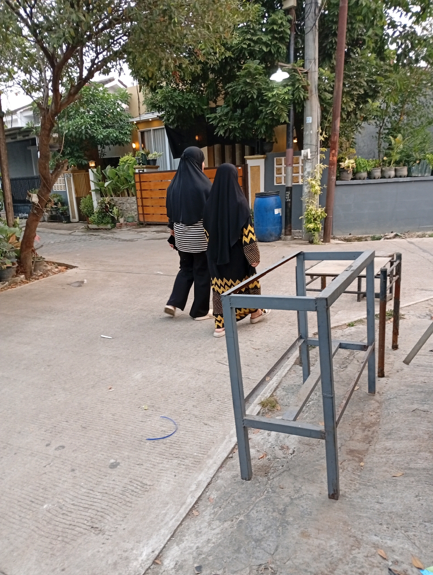 In an outdoor scene, two individuals wearing black hijabs are walking away from the camera on a paved walkway. The individual on the left wears a black hijab and a striped shirt with black pants. The individual on the right wears a black hijab and a black garment with a yellow zig-zag pattern. A blue barrel is visible behind them, along with a metal structure that appears to be a bench or table frame. The surrounding environment includes trees, potted plants, and a grey wall. The setting is a street or pathway in the city of Caringin, Indonesia, indicated by the context provided.