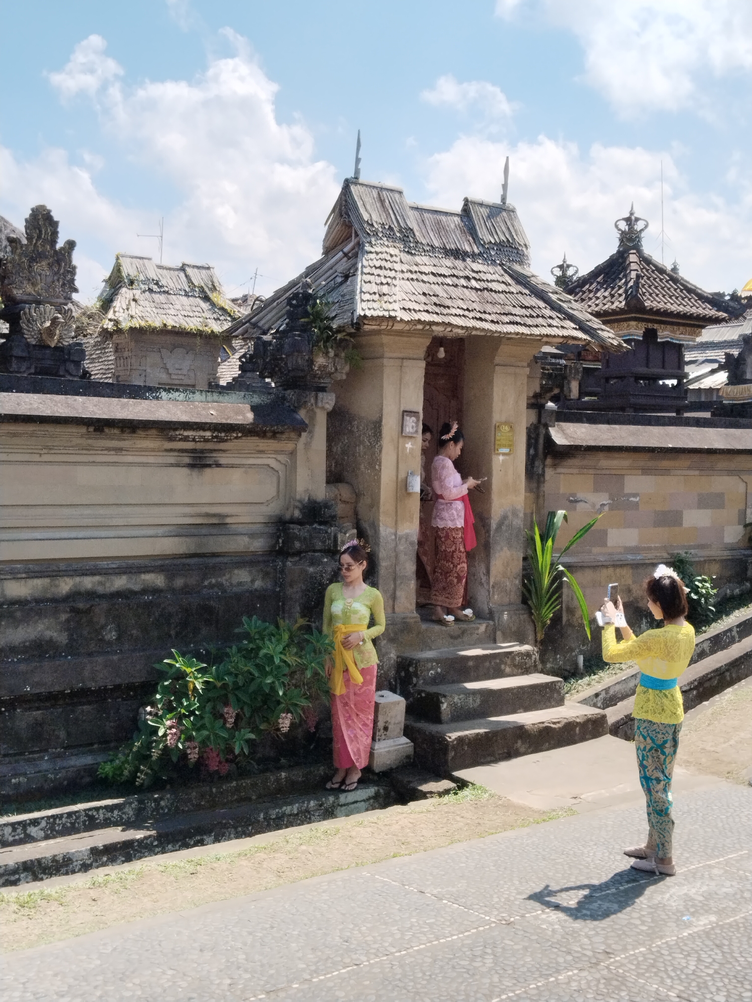 In the city of Bangli, Indonesia, on a sunny day with scattered clouds, three women are present in front of a traditional Balinese temple. The temple features a stone wall with decorative carvings and a thatched roof structure. One woman, wearing a yellow top and patterned pants, is taking a photograph of the other two women with her smartphone. The other two women are standing inside an entryway of the temple, both wearing traditional Balinese attire. One is in a pink and red outfit, and the other is in a pink and maroon outfit. Another woman, dressed in a yellow top and pink skirt, is standing to the left of the temple entrance, also posing. The ground in front of the temple is paved with stones, and there is greenery around the entrance.