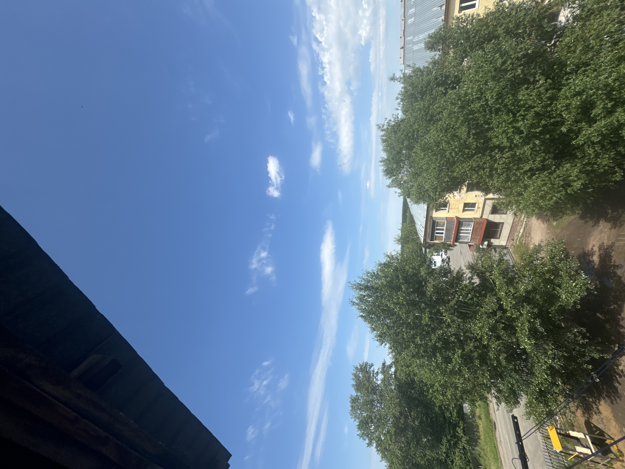 The image is a wide-angle shot taken from a high vantage point, looking up at a bright blue sky with scattered white clouds. In the foreground, a dark wooden overhang partially obscures the view. Below the sky, a dense canopy of green trees dominates the middle ground, with a small apartment building with yellow and brown siding visible between them. The building has multiple windows and balconies. Beyond the trees, a road and some other structures can be seen. The overall scene suggests an outdoor environment, possibly a residential area in a city. The date stamp indicates the image was taken on July 28, 2022, at 11:48 AM.
