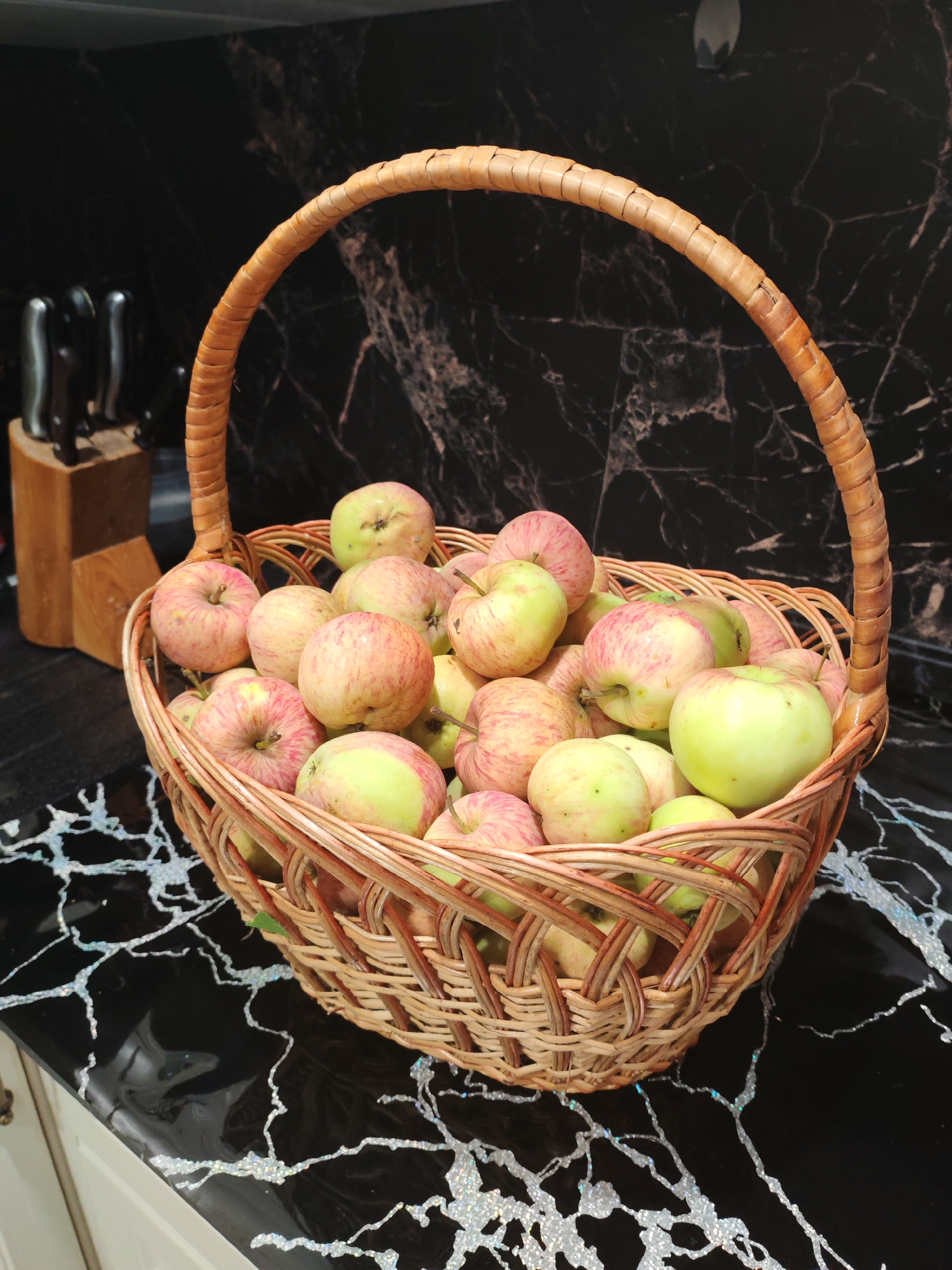 A close-up, eye-level shot shows a woven basket filled with apples on a black countertop with white, marble-like veins. The basket has a curved handle made of interwoven wicker. The apples are a mix of green and red-streaked varieties. In the background, a knife block with several knives is visible against a dark, patterned wall. The scene appears to be indoors, likely a kitchen, and is devoid of any people or specific city identifiable markers.