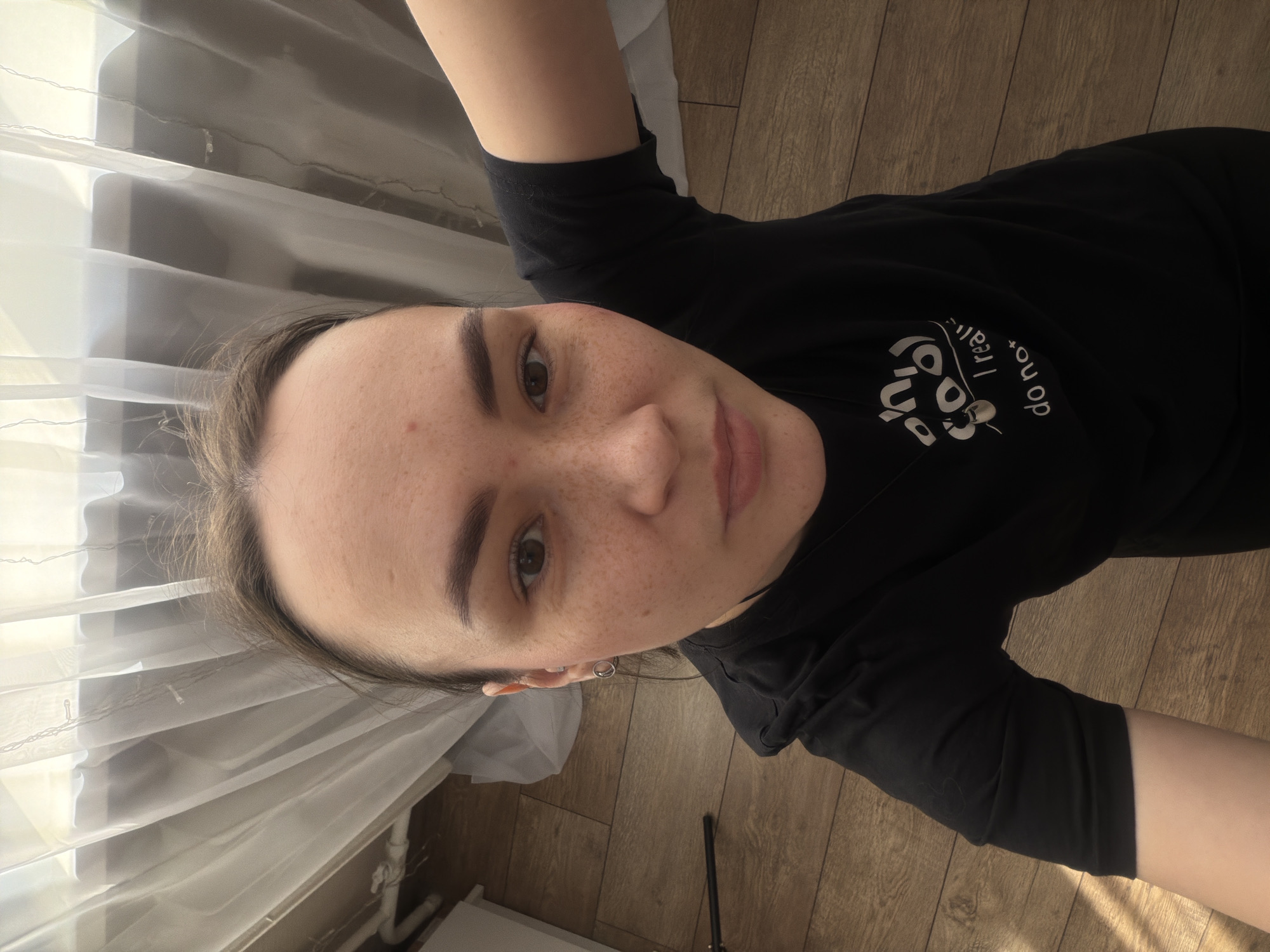 A high-angle selfie photograph depicts a female subject indoors in what is identified as the city of Chaikovsky, Russia. The subject, positioned centrally, has brown hair, dark brown eyes, and a fair complexion with numerous freckles across her nose and cheeks. Small red blemishes are visible on her forehead. She wears a black short-sleeved t-shirt with white text partially visible on the chest, reading "oh so I really do not". A small, silver-colored hoop earring is present in her left ear. Her gaze is directed towards the camera. The background consists of a window covered by sheer, light-colored curtains, with ambient light filtering through. Below the curtains and to the left, a white pipe or conduit is visible. The flooring is a light brown wood-grain laminate. A black rod-like object lies on the floor to the left of the subject.