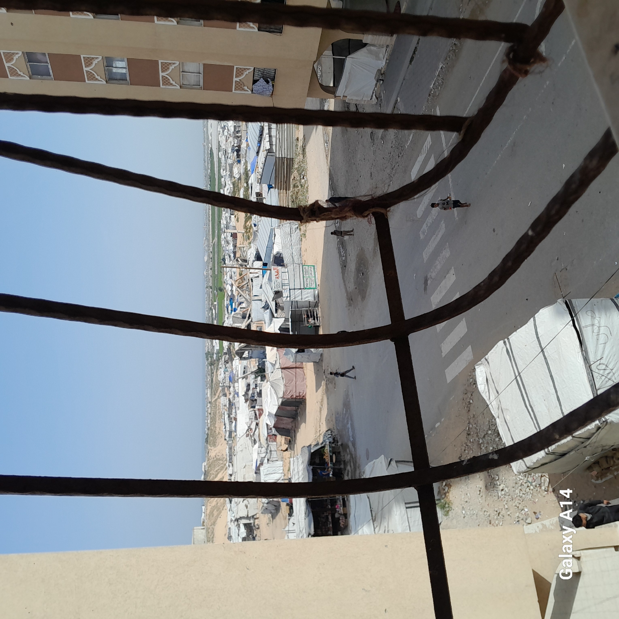 A high-angle view, likely taken from a balcony or window, captures a scene overlooking a densely packed settlement. In the foreground, a sturdy metal railing frames the view. To the right, a multi-story building with tan and brown sections features windows, some adorned with decorative white patterns. Laundry hangs from one of these windows.
A dusty road or street runs through the midground, with faint white crosswalk markings visible. Several individuals are present on the road; one person is clearly seen walking near the crosswalk, wearing a white t-shirt with "GM" or "OM" printed on it and dark shorts. Another figure in darker attire is further down the road.
To the left, a sprawling informal settlement extends into the distance, characterized by numerous makeshift structures, tents, and shelters constructed from tarpaulins and various materials. Within the settlement, a green signboard with Arabic text is visible, indicating local businesses or services such as a "barber shop" (صالون حلاقة), "vegetable shop" (محل خضراوات), and "restaurant" (مطعم). Beyond the camp, patches of green agricultural fields stretch towards the horizon under a clear, light blue sky.
The weather is bright and sunny, indicative of daytime. The overall scene suggests a refugee or internally displaced persons (IDP) camp, with daily life activities, including people walking and commerce, taking place amidst the temporary housing. A "Galaxy A14" watermark is present in the bottom right corner of the original image.