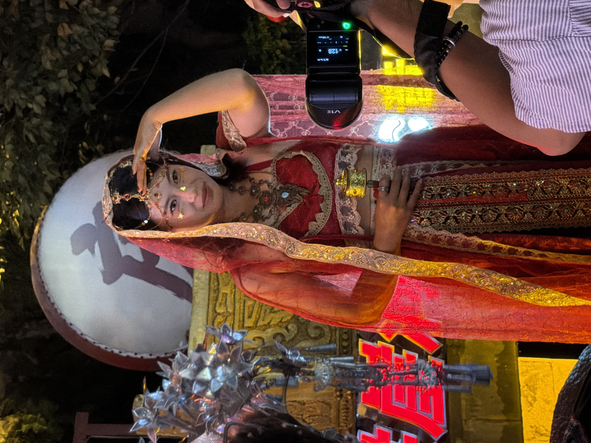 The image captures a person dressed in traditional attire, possibly a dancer or performer, at what appears to be an outdoor cultural event or festival. The individual wears a vibrant red and gold costume, adorned with intricate patterns and jewelry. A sheer red veil with golden trim covers their head and shoulders. The person is looking towards the right with their right hand raised to their forehead, possibly shielding their eyes from a light source or acknowledging someone. In their left hand, they hold a small, ornate golden object that resembles a prayer wheel or decorative bell.
Behind the performer, a large drum with a red rim and a white surface bearing a dark, stylized Chinese character is visible. To the left of the performer, there are metallic, decorative elements with a spiky, floral design, hinting at a float or stage decoration. In the lower left corner of the image, bright red, illuminated Chinese characters are visible, possibly part of signage for the event. The background consists of dark foliage and the warm glow of lights, suggesting an evening or night setting. A camera flash unit is prominently positioned in the lower right quadrant of the image, indicating that the scene is being photographed, likely by a spectator or professional. The presence of the camera and the festive atmosphere suggest the location is a public space within the city of Yuzhong District, China, during a celebratory event.