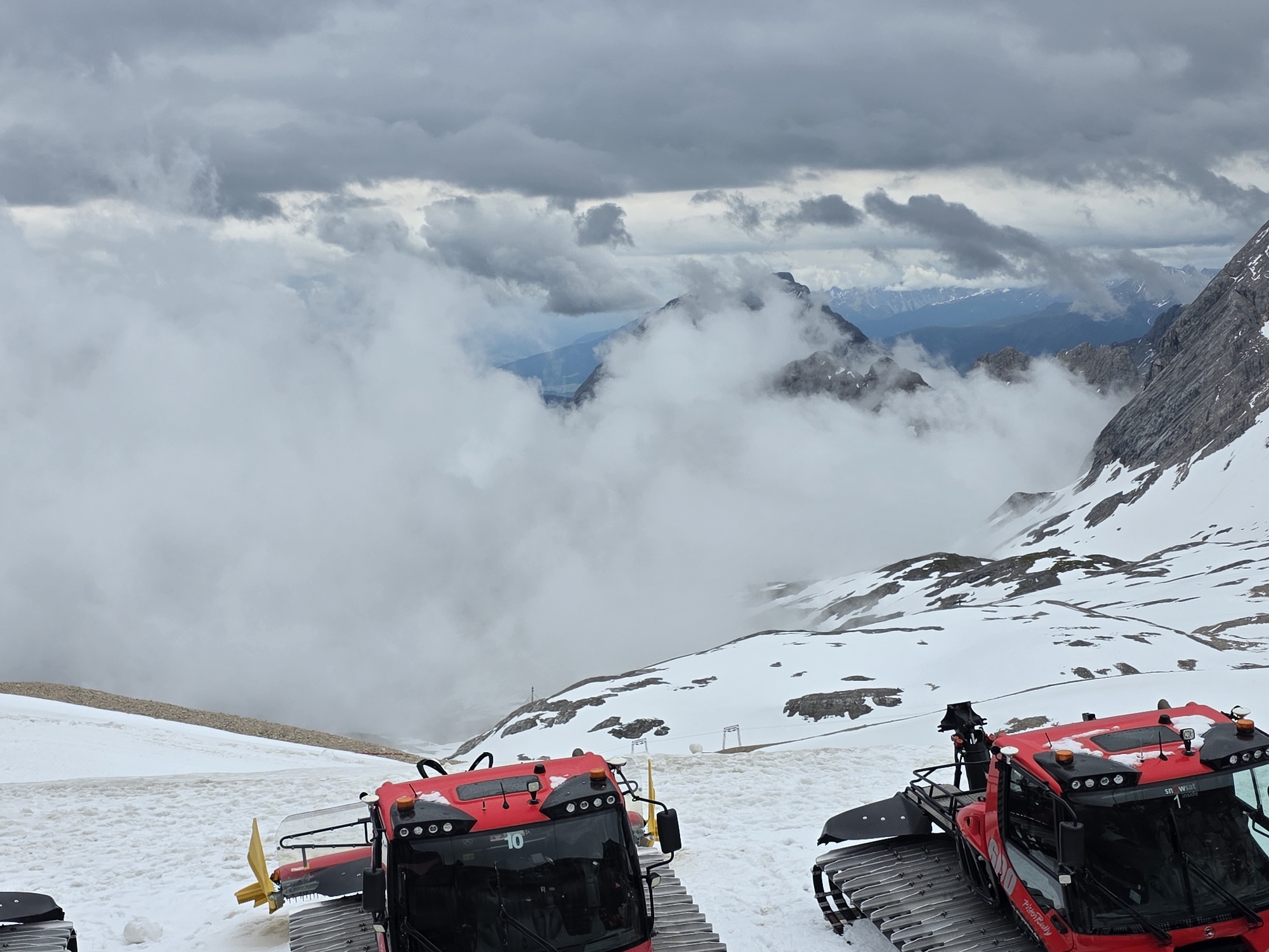 This image captures a high-altitude alpine environment, likely within the Schneeferner glacier area near Garmisch-Partenkirchen, Germany, on an overcast day. In the immediate foreground, two red PistenBully snow groomers are stationary on a snow-covered slope. The groomer on the left, identified with the number "10" on its front windshield, presents a yellow blade attachment. The second groomer on the right displays "PistenBully" branding on its side and "snowsat" with the number "1" on its cabin roof. The ground beneath the vehicles consists of patchy snow mixed with exposed brown earth or rock, suggesting areas of melt.
The midground features a gently sloping snowfield punctuated by darker, exposed rock formations. A distinct, horizontally oriented band of brown earth is visible across the upper-left midground, contrasting with the snow. The background is characterized by a dense layer of white clouds and fog that obscures the lower and mid-sections of rugged mountain peaks. Above, a heavily overcast sky with dark grey storm clouds dominates, while glimpses of distant, blue-tinged mountains are visible through breaks in the cloud cover. No human figures are present in the scene.