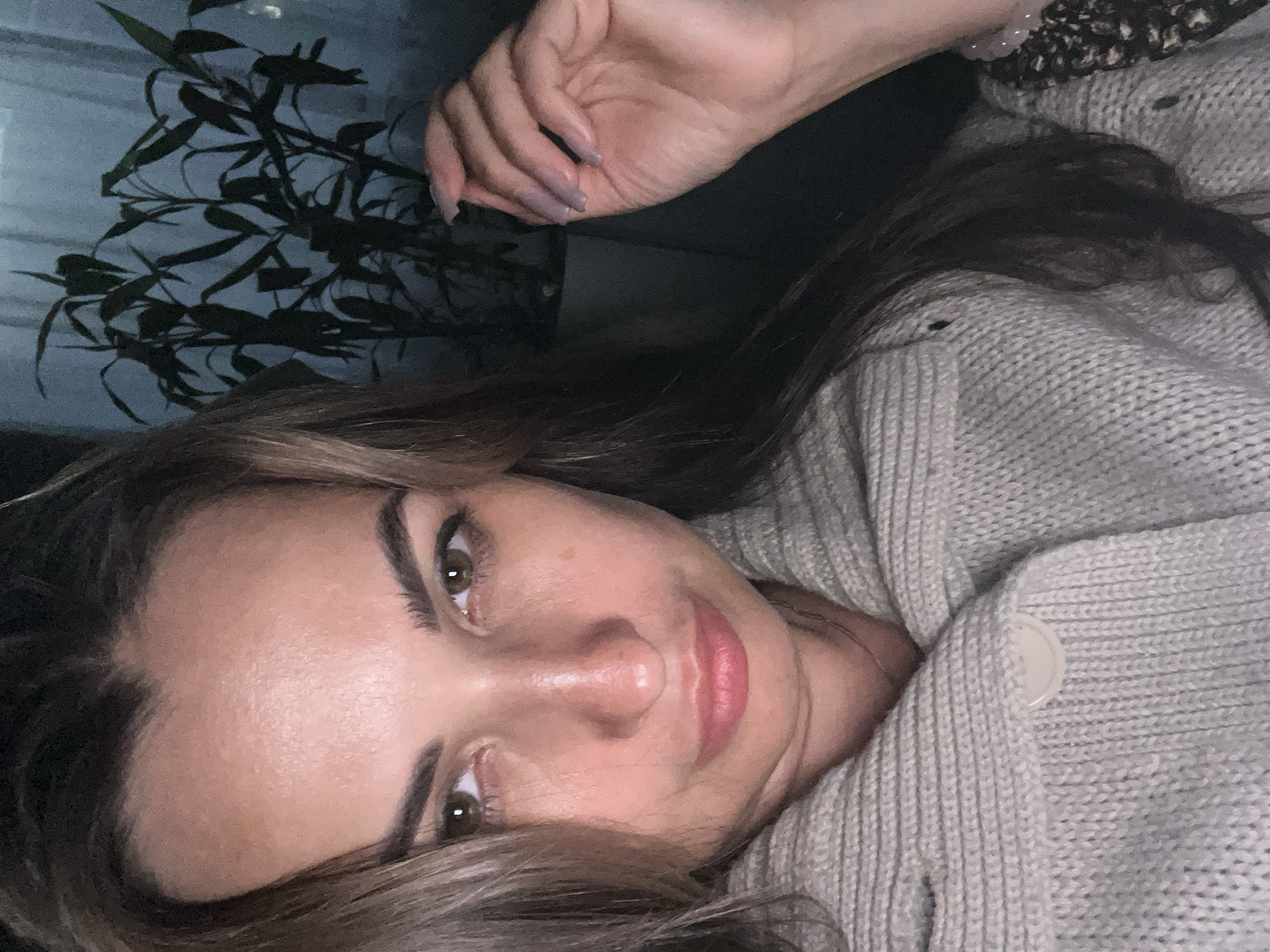 A woman with light brown hair and brown eyes is looking at the camera. She is wearing a light gray knitted cardigan. Behind her, a dark green plant is visible against a dark background with a hint of blue. A hand with light purple nails is resting near the plant. The overall lighting is dim. The location is Unknown, Unknown.