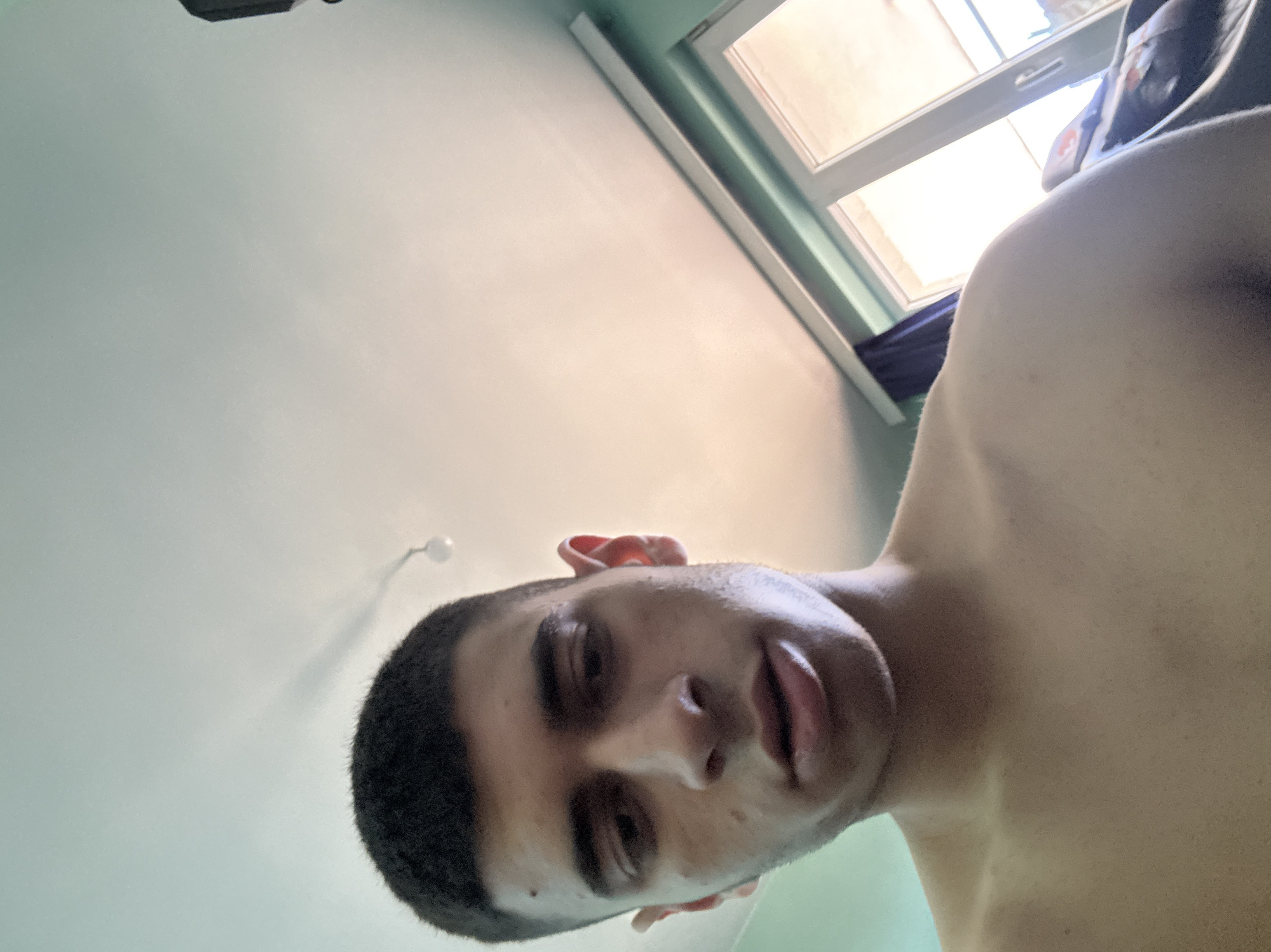 A close-up, low-angle shot reveals a man with dark hair and light skin. He is shirtless, his head tilted downward towards the camera. The background consists of a light green wall and a window with a white frame. A bare bulb ceiling fixture is visible. There is no evidence to suggest that this photo was taken in Sofia, Bulgaria.