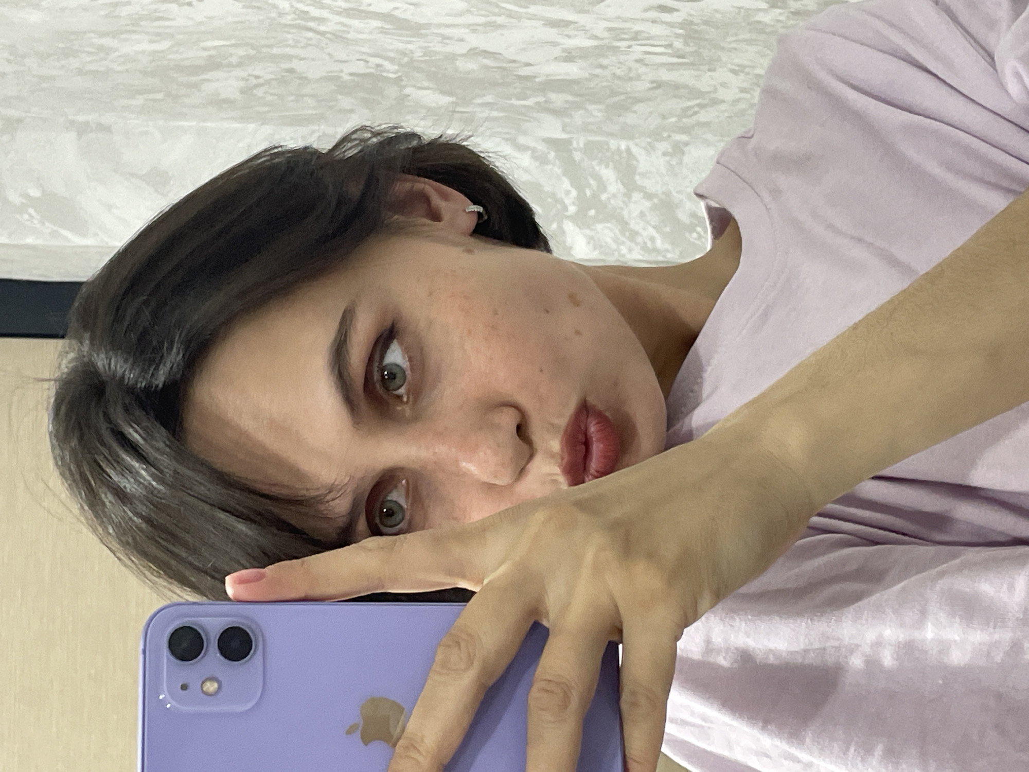 A woman is taking a selfie with a purple iPhone 11. The background appears to be a white wall. She is wearing a light purple t-shirt and has dark brown hair. She is looking at the camera with her head tilted to the left. There is no indication of the location being Izhevsk, Russia.