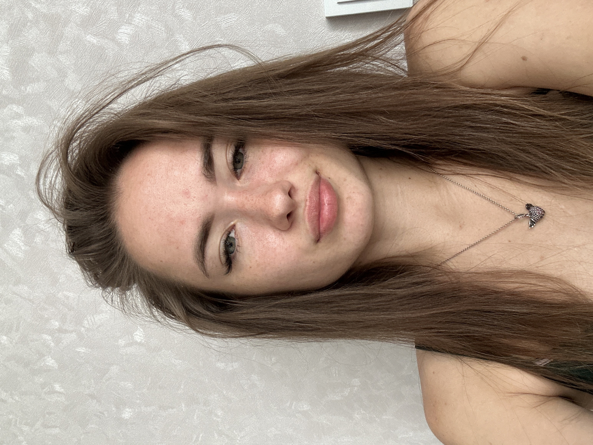 A young woman with long brown hair is pictured from the shoulders up. She is looking directly at the camera with a neutral expression. Her skin is fair with some freckles and minor blemishes. She is wearing a dark-colored top and a silver necklace with a small pendant. The background is a textured, light-colored wall.