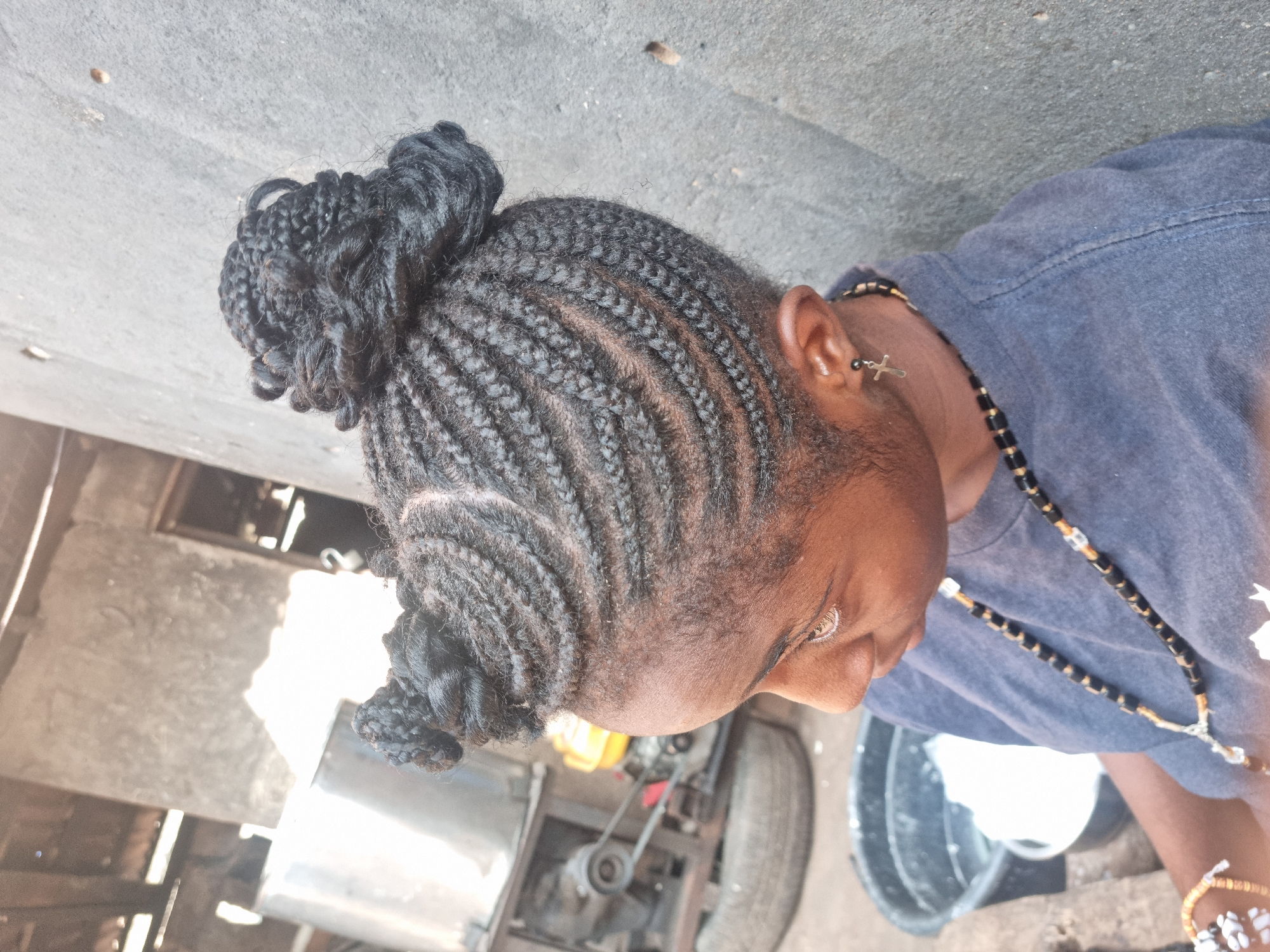 This media file captures a close-up, slightly high-angle view of a person with dark skin, focusing on their head and upper body. The person's hair is styled in neat, intricate cornrows that lead into two buns: a larger one at the crown of the head and a smaller one towards the back of the neck. The braids expose lines of scalp.
The individual is wearing a casual blue or gray short-sleeved t-shirt. Visible accessories include a silver cross-shaped earring in their right ear and a long necklace made of black and light-colored beads. A multi-colored bracelet is also partially visible on their right wrist. The person's gaze is directed downwards and slightly to the right, suggesting they are focused on an activity or object out of frame. Their expression is neutral and concentrated.
The setting appears to be outdoors during bright daylight, indicated by strong lighting and shadows. The background is somewhat cluttered and industrial. To the lower left, mechanical components like a motor, pulleys, and belts, possibly belonging to an engine or generator, are visible. A large tire is also present in the mid-ground on the right. A dark bucket is partially seen on the far right. The ground beneath them seems to be a light-colored, possibly concrete or dusty paved surface. The scene suggests an urban or semi-urban outdoor environment, potentially a workshop or a casual outdoor work area in Onitsha, Nigeria. No visible text is clearly legible in the image.