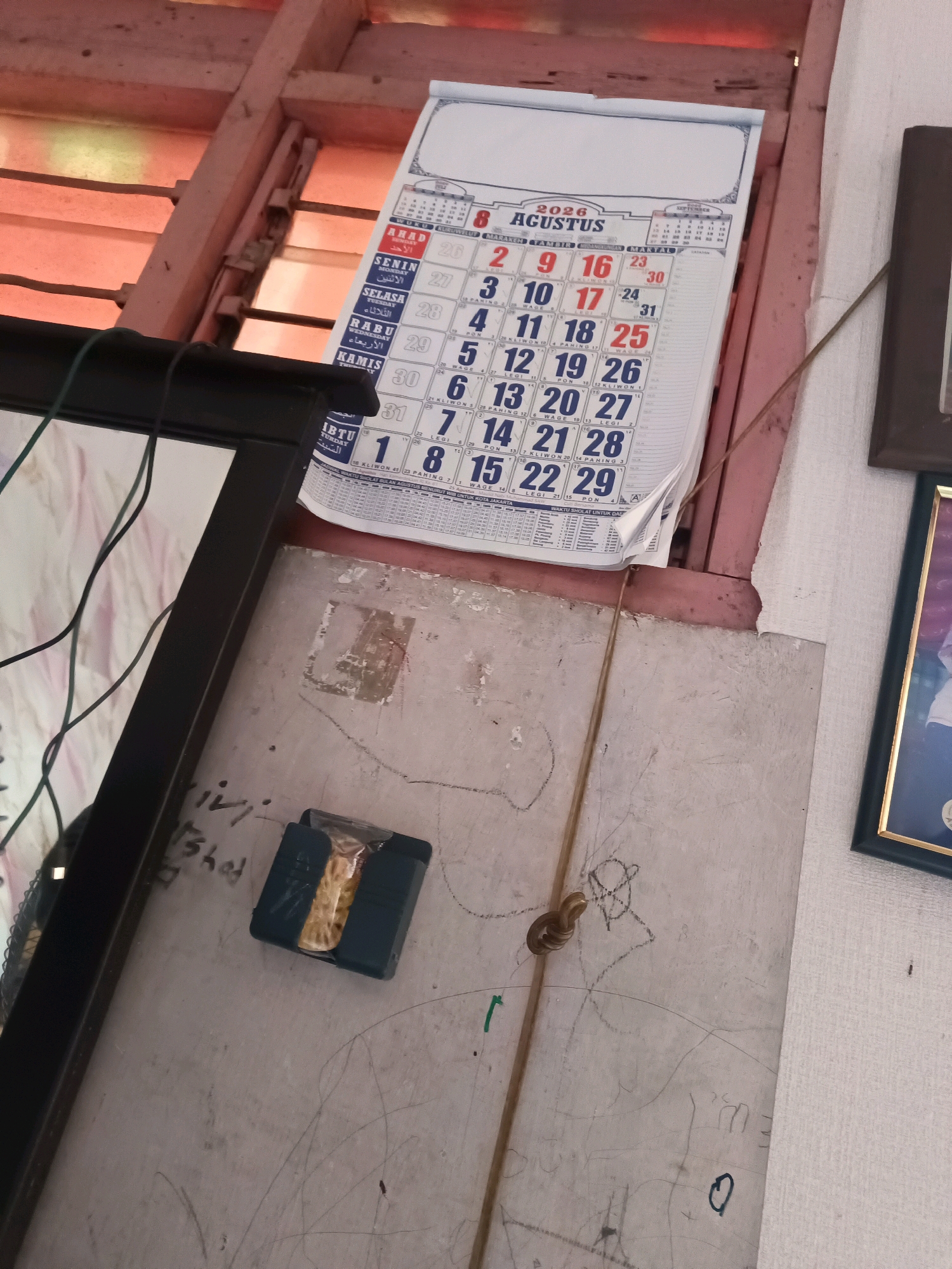 The image depicts a close-up interior view of a wall with a calendar. The calendar, dated August 2026, hangs on a light-colored wall that shows signs of wear and has various markings and scribbles, some appearing to be children's drawings. To the left of the calendar, a black framed picture is partially visible. Below the calendar, a small dark blue holder contains what appears to be a pack of small items, possibly snacks or candies. A thin brown cord runs vertically down the wall, attached to a small metal hook. The background shows wooden beams and a window with translucent blinds, suggesting an indoor environment. The overall impression is of a lived-in space.