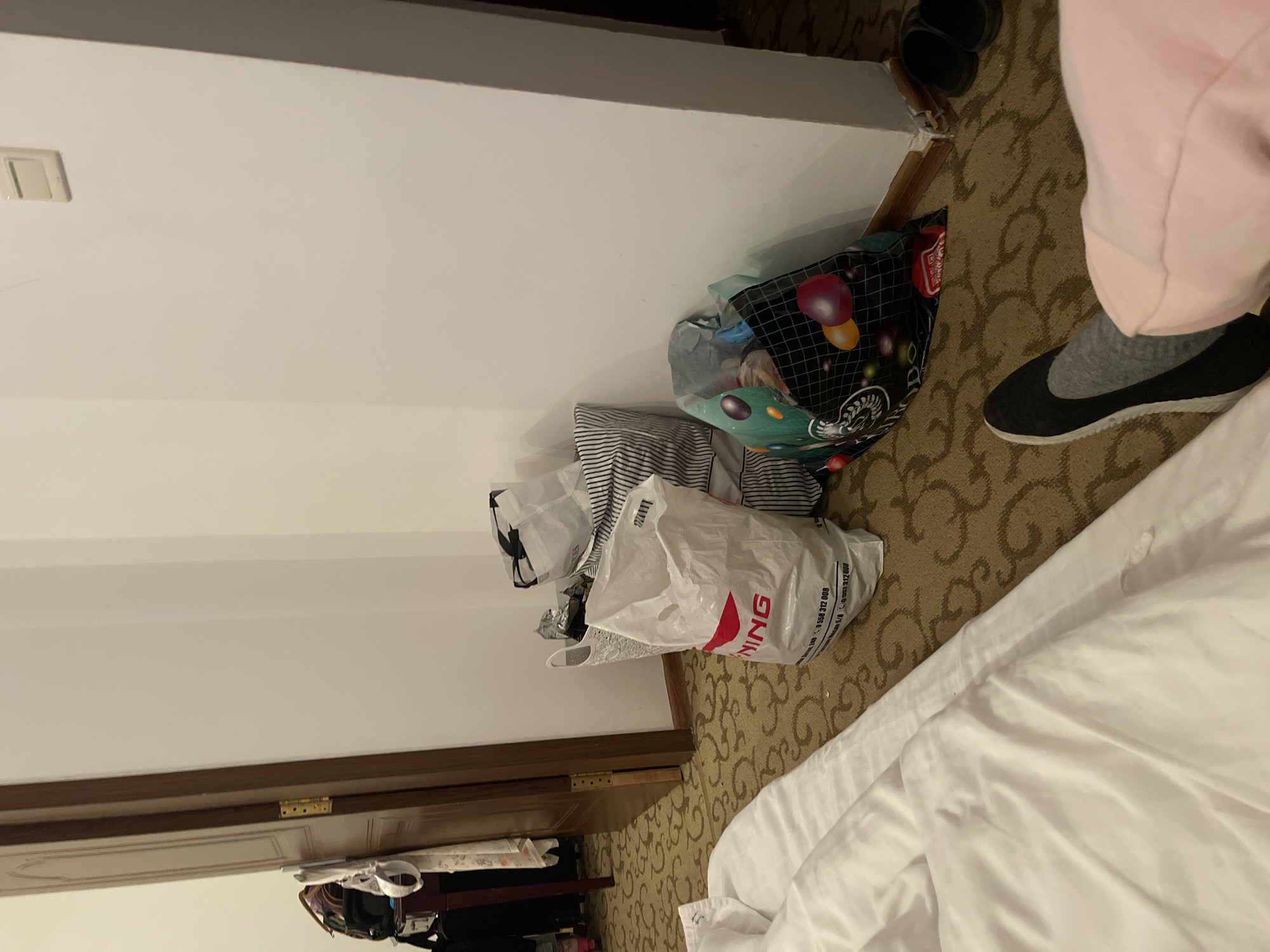 The image depicts an indoor scene, likely a hotel room in Ornek, Kyrgyzstan, based on the provided context. A corner of a large white bed with rumpled sheets dominates the lower left of the frame. The rest of the room features a patterned beige carpet and a white wall. Several shopping bags are gathered near the wall. One is a large white plastic bag with red text reading "NING" and contact information. Next to it is a black and white striped bag, partially obscured by a white woven basket holding some items. Another bag, black with a colorful geometric pattern, is placed on the floor. A wooden door is slightly ajar on the left side of the image, revealing a glimpse of furniture and possibly other bags. A light switch is visible on the upper right portion of the wall. In the lower right corner, a pair of black slippers rests on the carpet, and a person's legs, clad in pink loungewear and a grey sock with a black sneaker, are visible, suggesting someone is seated or lying on the bed. The overall impression is one of a temporary living space with items that have been recently unpacked or are prepared for departure.