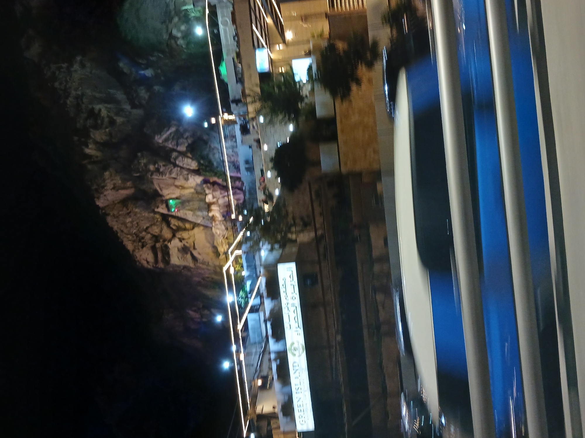 The image captures a nighttime scene with a prominent rocky hillside illuminated by artificial lights. Below the hillside, a modern building structure is visible, featuring white and light-colored walls and a facade adorned with horizontal strips of warm-toned lights. A large sign with Arabic text and the English words "GREEN ISLAND" is mounted on the building. The building appears to be a restaurant or commercial establishment. In the foreground, a portion of a vehicle, possibly a bus or van, with blue and white coloring is in motion, indicated by motion blur. The environment suggests an urban setting, possibly a scenic overlook or a recreational area, given the illuminated rocky terrain and the presence of a commercial establishment. The overall impression is one of a vibrant nighttime urban landscape with a blend of natural and man-made elements.