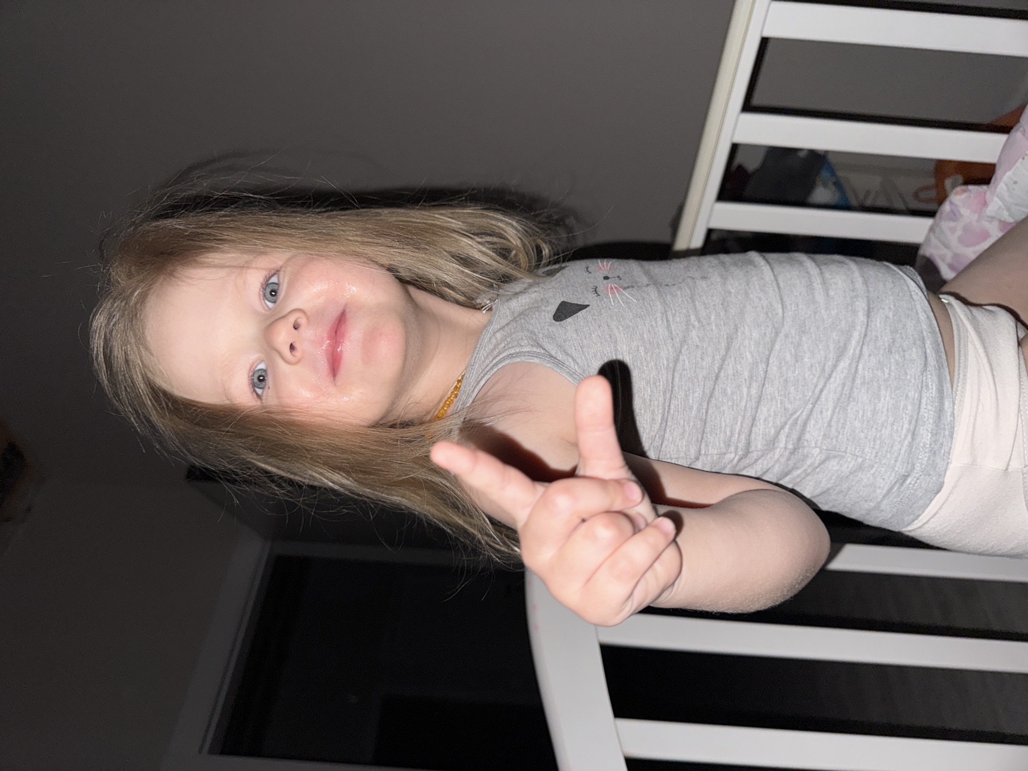 A young child with blonde hair and blue eyes is lying in a white crib. The child is wearing a light gray shirt with a cat design on the chest and light-colored bottoms. The child is making a peace sign with their right hand. The room appears to be dimly lit, with a dark wall and a window visible in the background. It is nighttime. No information related to the location of Gornyi (Dvurechenskoe S/P), Russia, is visible.