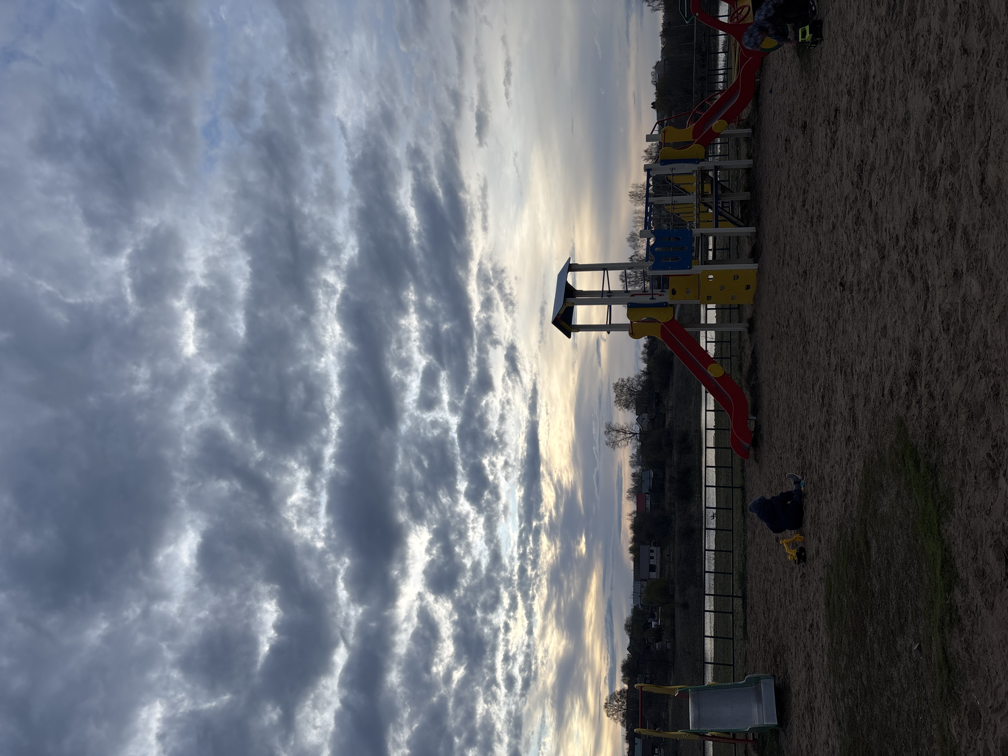 This image captures a playground scene under a dynamic, cloudy sky, likely during late afternoon or early evening in Foky, Russia.
The primary subject is a colorful playground structure situated on sandy ground. It features multiple platforms, red and silver slides, and climbing panels in yellow and blue. To the right of the frame, a child, dressed in dark clothing, is seated on the sand, engrossed in playing with a yellow toy excavator.
Behind the playground, a dark metal fence separates the play area from a body of water, which appears to be a river or lake, reflecting the low-lying light. On the distant bank, a line of trees and scattered houses are visible against the horizon.
The sky is a notable feature, dominated by extensive, wavy cloud formations, possibly altocumulus undulatus. Patches of brighter, lighter clouds and clear sky are interspersed, allowing sunlight to break through and illuminate parts of the scene with a soft, diffused glow. The lighting suggests the sun is low, casting a gentle light across the landscape. The overall atmosphere is tranquil and focused on the child's play.
No visible text is present in the image.