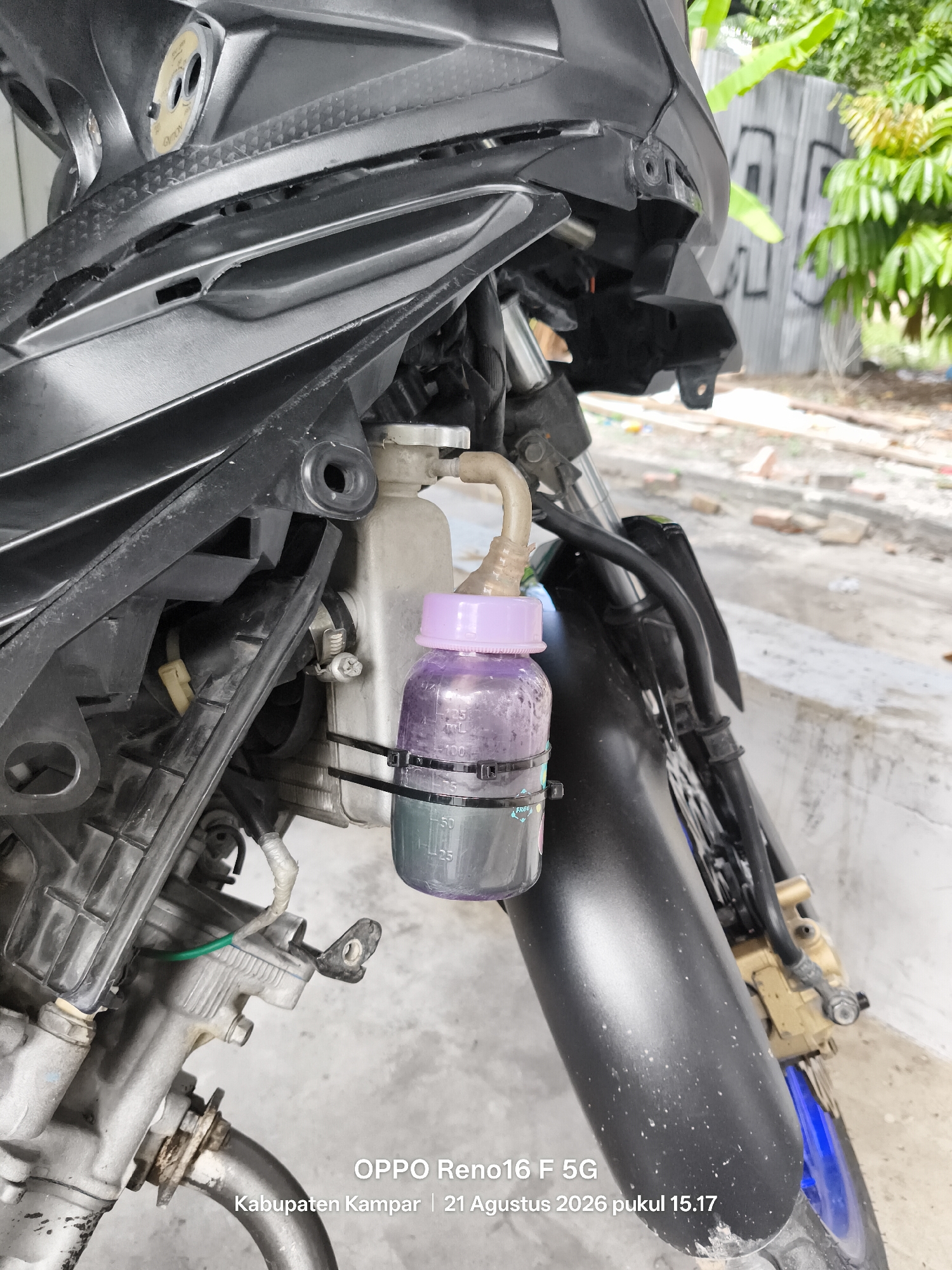 The image shows a close-up of a modified motorcycle's front section. A small plastic bottle, filled with a purple liquid and capped with a light purple lid, is attached to the motorcycle's frame using black zip ties. A thin, flexible tube connects the bottle to a reservoir, which appears to be part of the motorcycle's cooling system. The motorcycle is black and has some silver and gold-colored mechanical parts visible. The background is blurred, showing a ground surface with some scattered debris, possibly dirt and small stones. Green foliage and a gray, graffiti-covered wall are visible in the distance. The timestamp on the image indicates it was taken on August 21, 2026, at 3:17 PM in Kabupaten Kampar, Indonesia. There are no people or any discernible actions taking place in the scene.