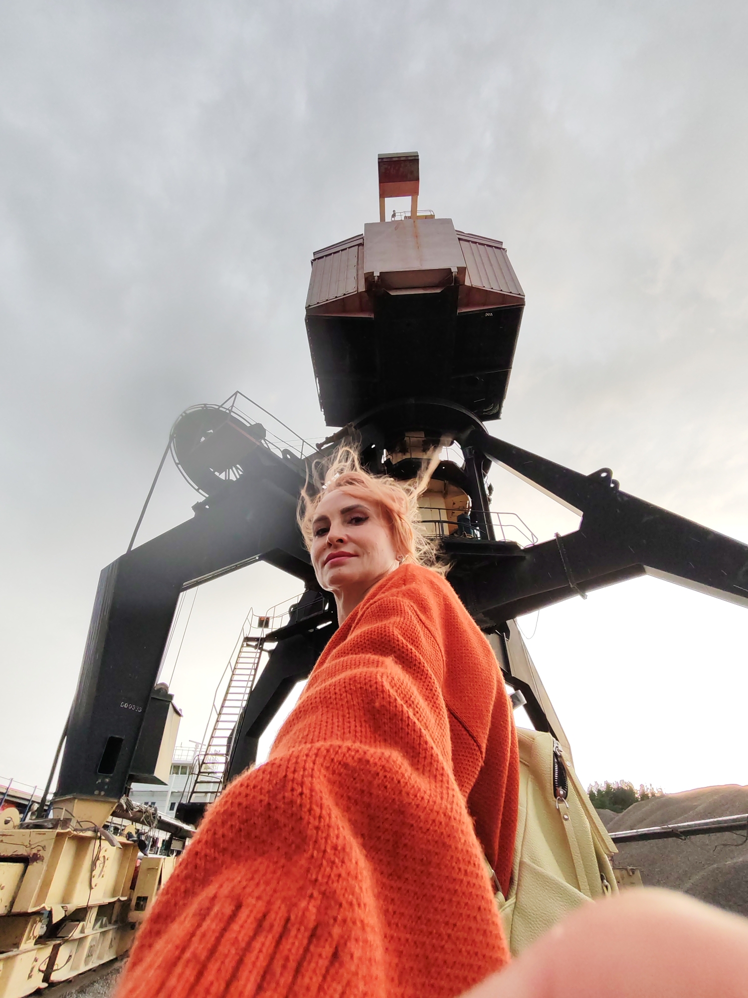 A low-angle, wide-shot, selfie-style photograph captures a woman with short, light-colored hair and makeup, looking towards the camera with a slight smile. She is wearing an oversized, bright orange knitted sweater and a light yellow backpack is visible over her left shoulder. She is positioned in front of a large industrial crane, which dominates the background. The crane is black and appears to be made of metal. Part of a ship or boat with a white superstructure is visible behind the crane, as well as a metal ladder ascending one of the crane's structures. The ground appears to be gravel. The sky is overcast and appears pale. The overall setting suggests an industrial port or quarry environment. The image was taken in the city of Shalsky, Russia.
