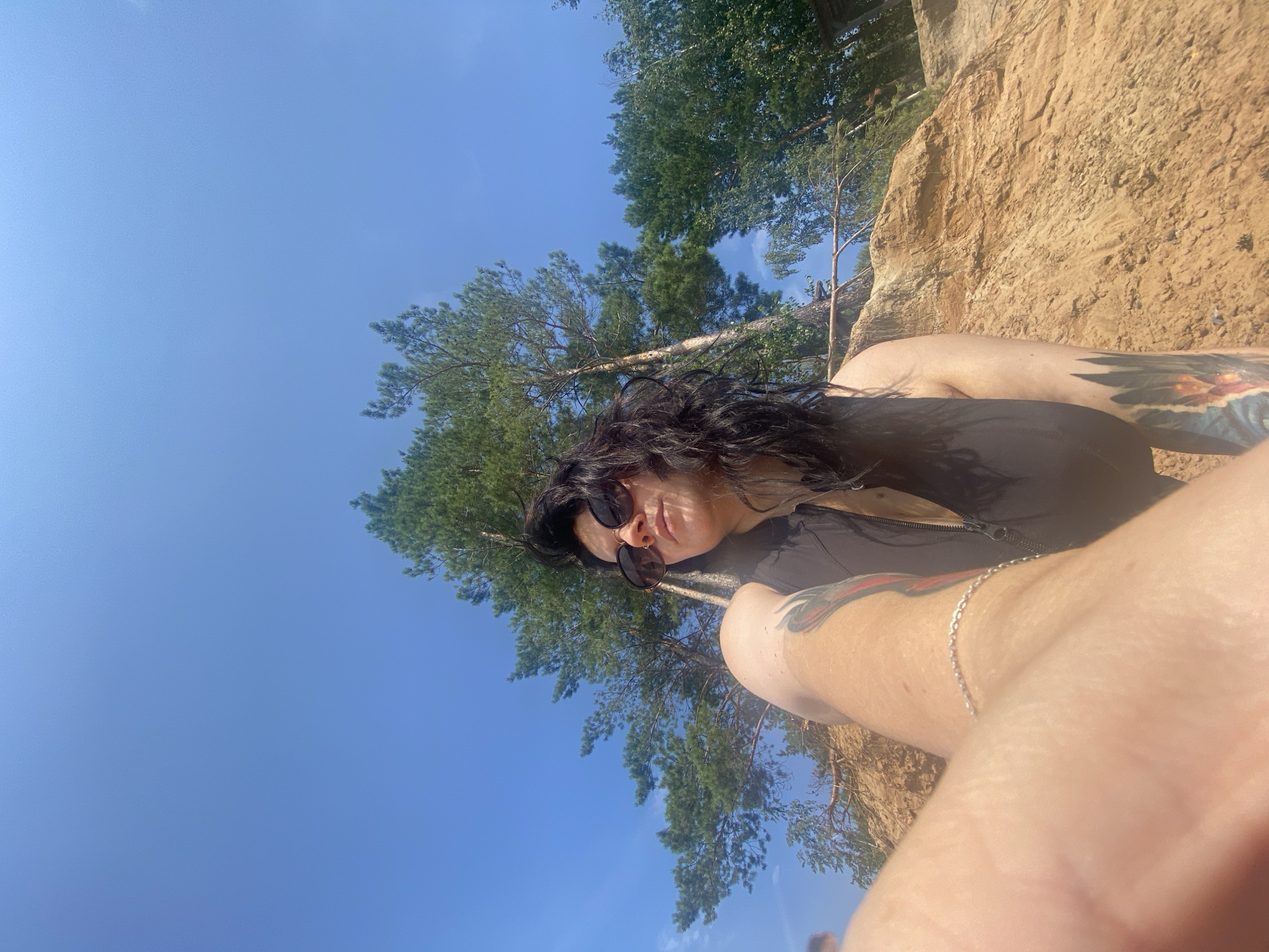 A woman with long, dark, wavy hair and sunglasses is sitting on a sandy slope under a bright blue sky. She is wearing a dark vest. Her left arm is visible in the foreground, showing a silver bracelet and tattoos on her upper arm and forearm. Behind her, tall pine trees and other greenery grow on the sandy embankment. The location is identified as Novyi, Russia. The scene appears to be a natural, outdoor setting.