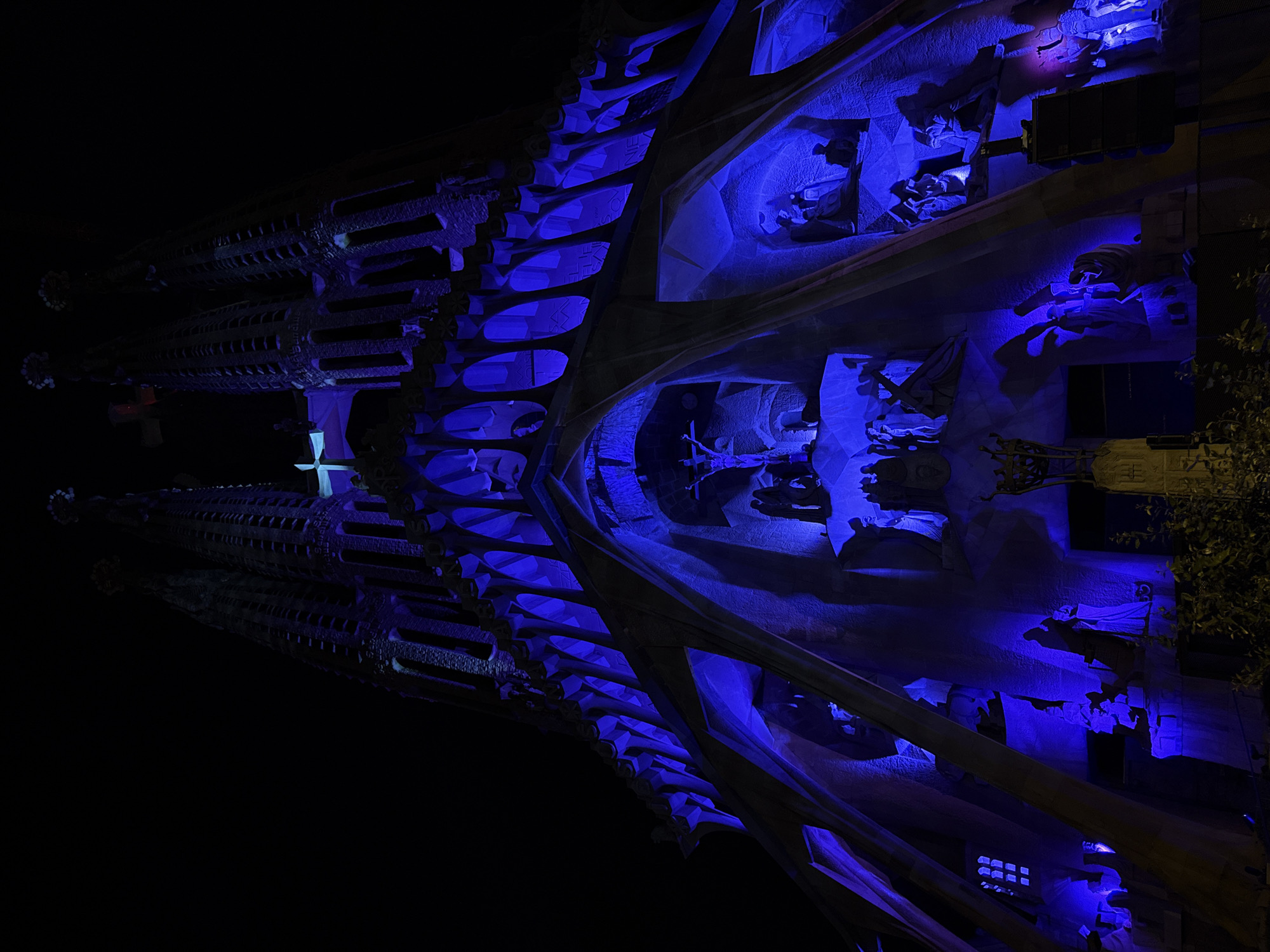 Here is a concise description of the image:
The image depicts a nighttime view of the Sagrada Familia basilica in Barcelona, Spain. The architectural details of the building are illuminated with blue lighting, highlighting the towers, sculpted facades, and intricate designs. The scene is dark, with the illuminated basilica standing out against the night sky. There are trees visible on the right side. The image is oriented sideways.