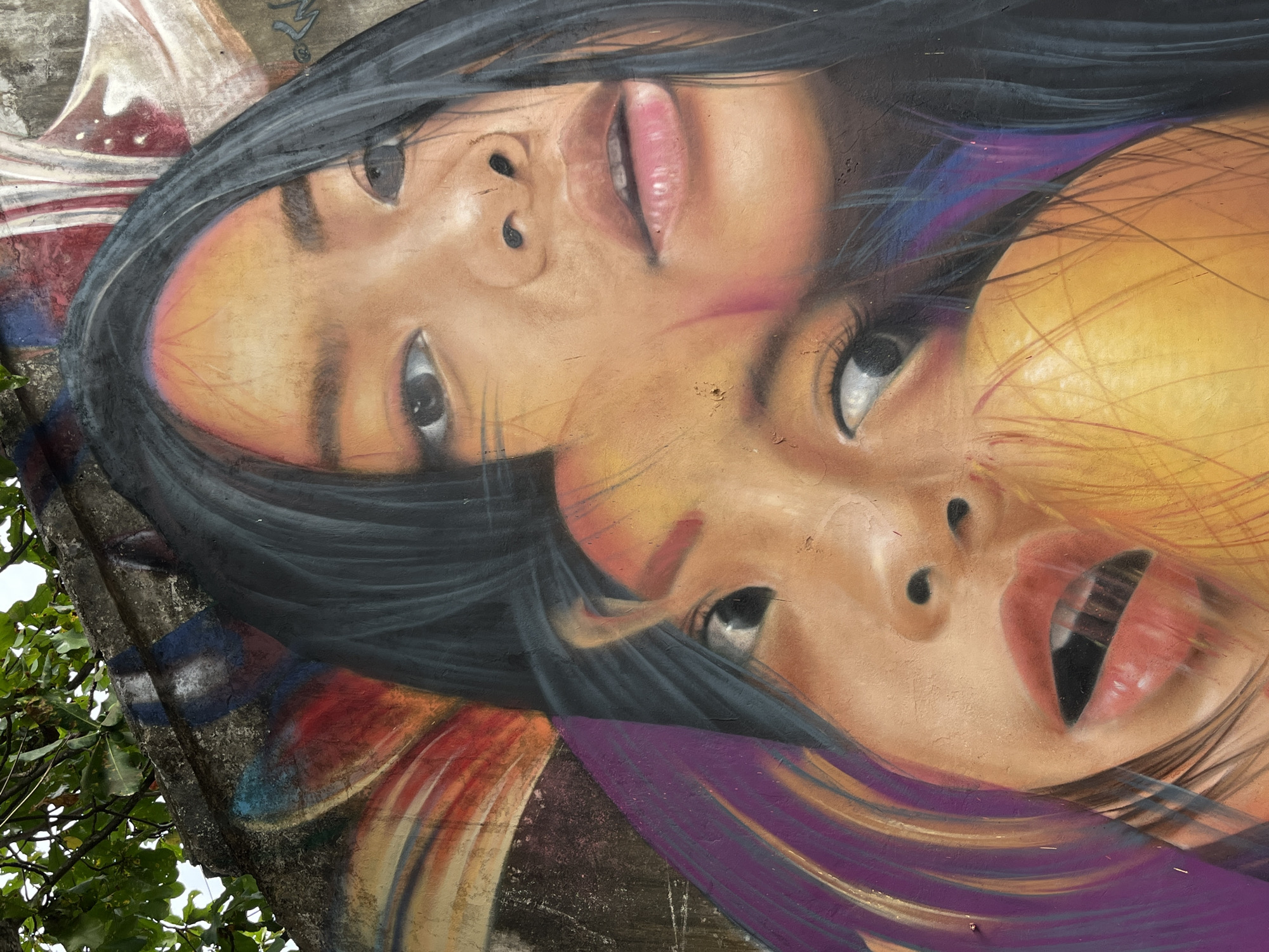 The image shows a close-up of a mural painted on a weathered concrete wall. The mural depicts two young girls, possibly sisters, their faces overlapping. The girl in the foreground has her mouth open, revealing dark teeth. The girl behind her has a softer expression, with her eyes looking slightly to the side. The mural is rendered in vibrant colors, with a focus on skin tones, dark hair, and colorful abstract elements surrounding the faces. The wall itself is textured and shows signs of age and weathering, with some peeling paint and graffiti visible in the background. Lush green foliage is visible in the upper left corner, suggesting an outdoor setting, possibly in a tropical environment. This artwork is located in the city of Kuta, Indonesia, where street art is a common sight, adorning many walls throughout the town.