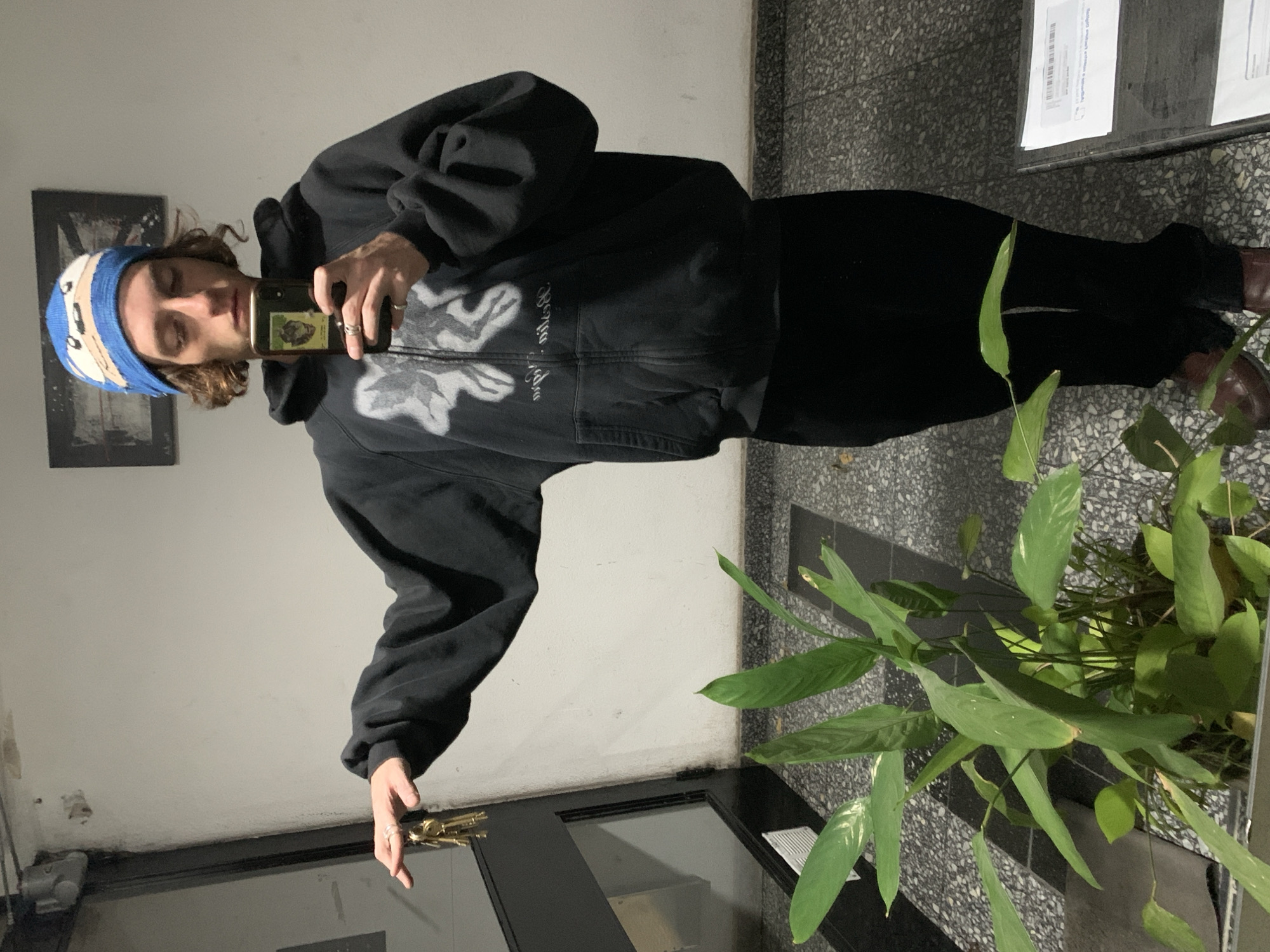 A young adult stands in a hallway, taking a selfie with a smartphone. The individual is wearing a blue and white striped beanie, a black hooded sweatshirt with a white graphic design on the front, black pants, and brown shoes. The setting appears to be an entryway with grey tiled floors and white walls. There is a houseplant in a grey container near the person's feet. Some mail and other papers are on the ground. The general environment suggests an apartment building hallway or entryway. The location is likely Buenos Aires, Argentina.