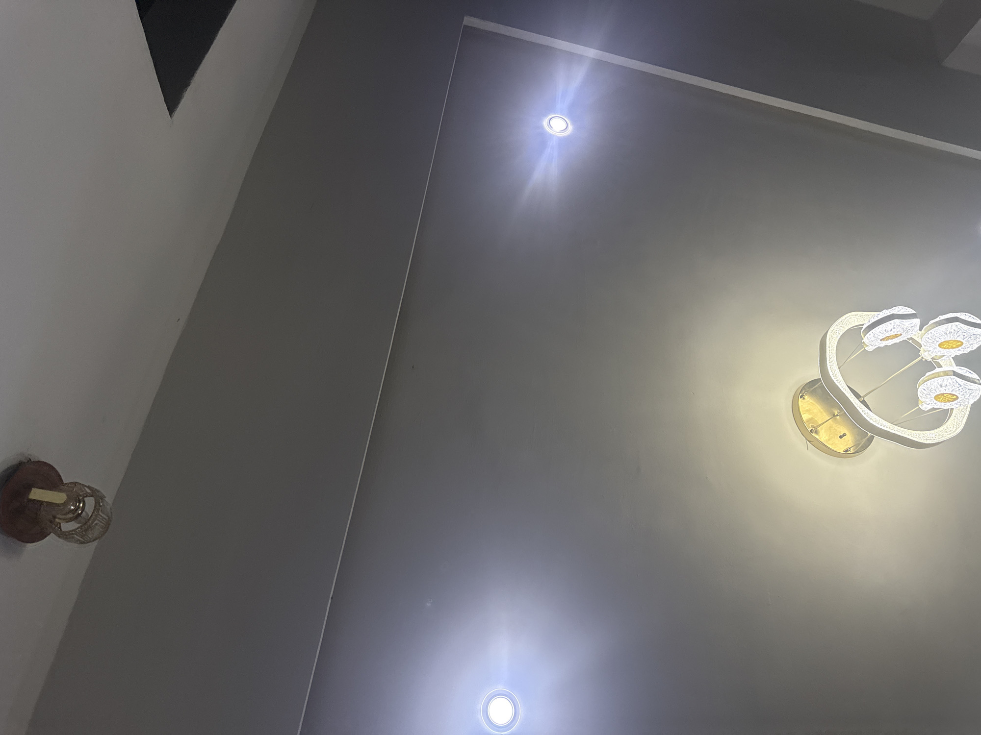 A high-angle shot focuses on the ceiling of an interior space. The ceiling is predominantly light gray, with three light sources visible: two recessed lights emit bright, focused beams, while a modern, multi-ringed chandelier provides a softer, more diffuse illumination. At the lower-left of the frame is a wall-mounted light fixture with a decorative shade. The image composition suggests the interior location is in Abuja, Nigeria due to the modern architectural design frequently found in residences there.