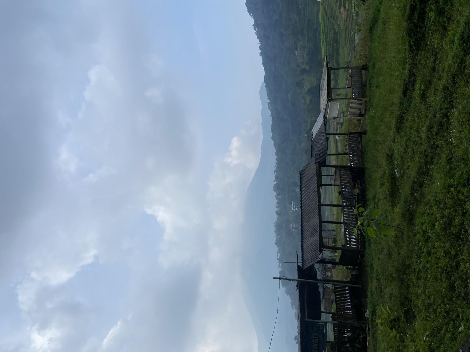 The image captures a scenic view in Tamiajeng, Indonesia, featuring a wooden shelter structure nestled among green vegetation. A mountain range is visible in the background, partially obscured by clouds. The sky is overcast. Adjacent to the shelter are terraced rice paddies. The photograph is oriented sideways.