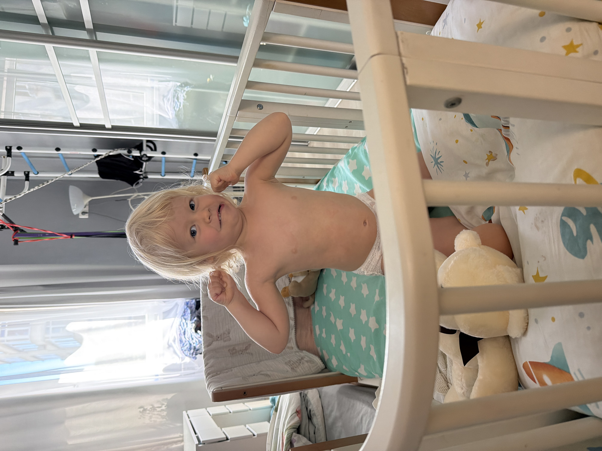 This media file captures a playful toddler with light blonde hair and fair skin, positioned inside a white wooden crib. The child, shirtless and wearing a white diaper, stands or kneels, looking directly at the viewer with a broad, happy smile. Both arms are raised and bent at the elbows, suggesting a playful flex or gesture.
The crib contains a mattress covered by a light teal sheet patterned with white stars, along with bedding featuring colorful space-themed illustrations of planets and stars. A cream-colored teddy bear with a black bow tie is visible nestled near the child.
The background shows a brightly lit interior, likely a child's room. A large window with a view of the outside provides ample daylight. Below the window, a white drying rack holds a dark piece of clothing, and various colorful cables are visible. A white lamp or light fixture hangs nearby, and a modular storage unit is partially seen to the lower left. The overall mood is cheerful, accentuated by the child's expression. The image itself is rotated 90 degrees clockwise, presenting the scene sideways. No text is visible in the frame. The location context is Sochi, Russia.
