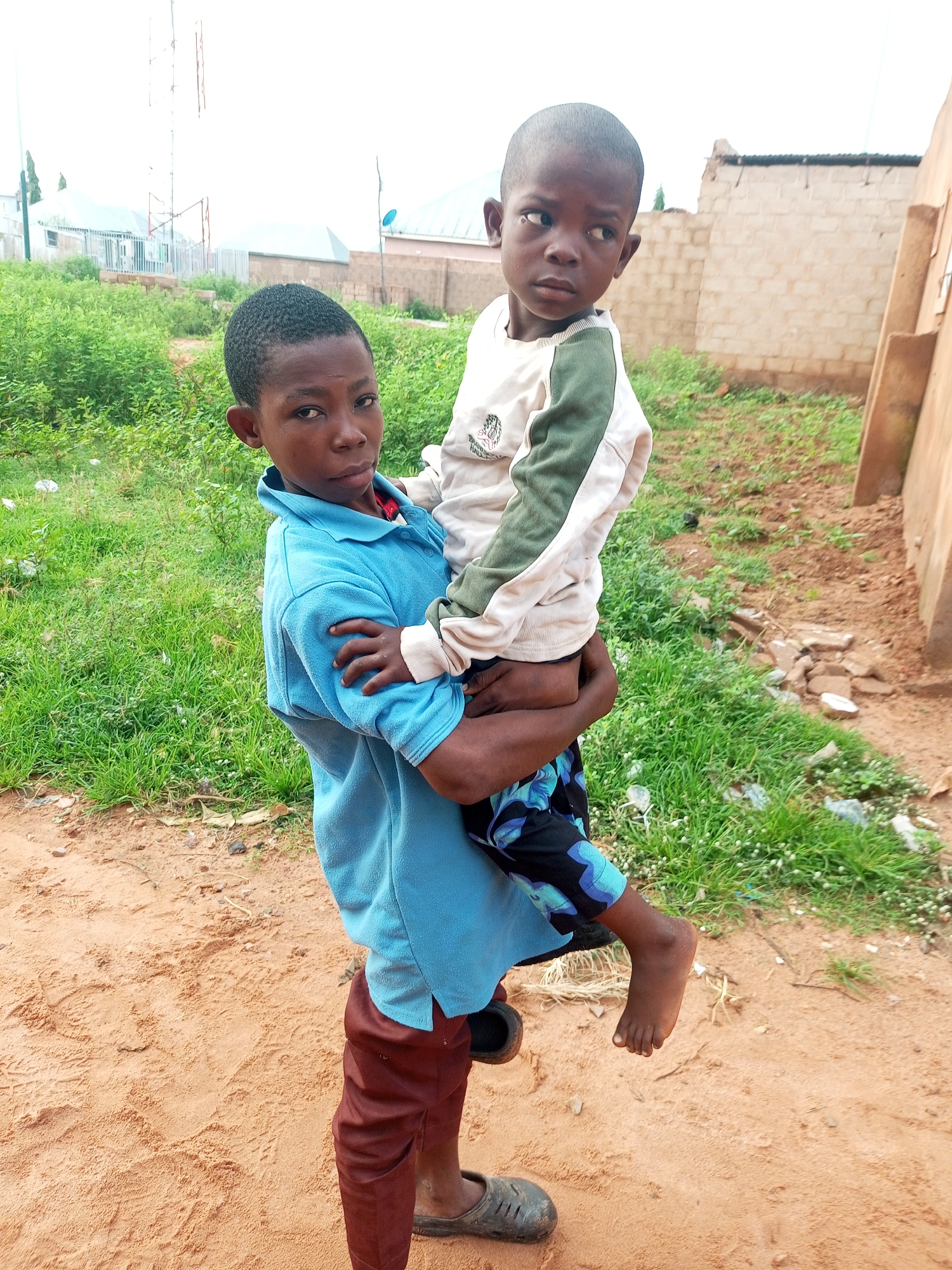 In Tempure, Nigeria, a young boy in a blue shirt and brown pants carries another boy in a cream-colored sweatshirt with green accents. The older boy faces the camera, his expression serious, while the younger boy looks to his left. They are in an outdoor environment with sandy ground and green vegetation. In the background, there are buildings, including one made of brick with a corrugated metal roof, and a tall tower. The overall scene appears to be a residential or undeveloped area within the city.