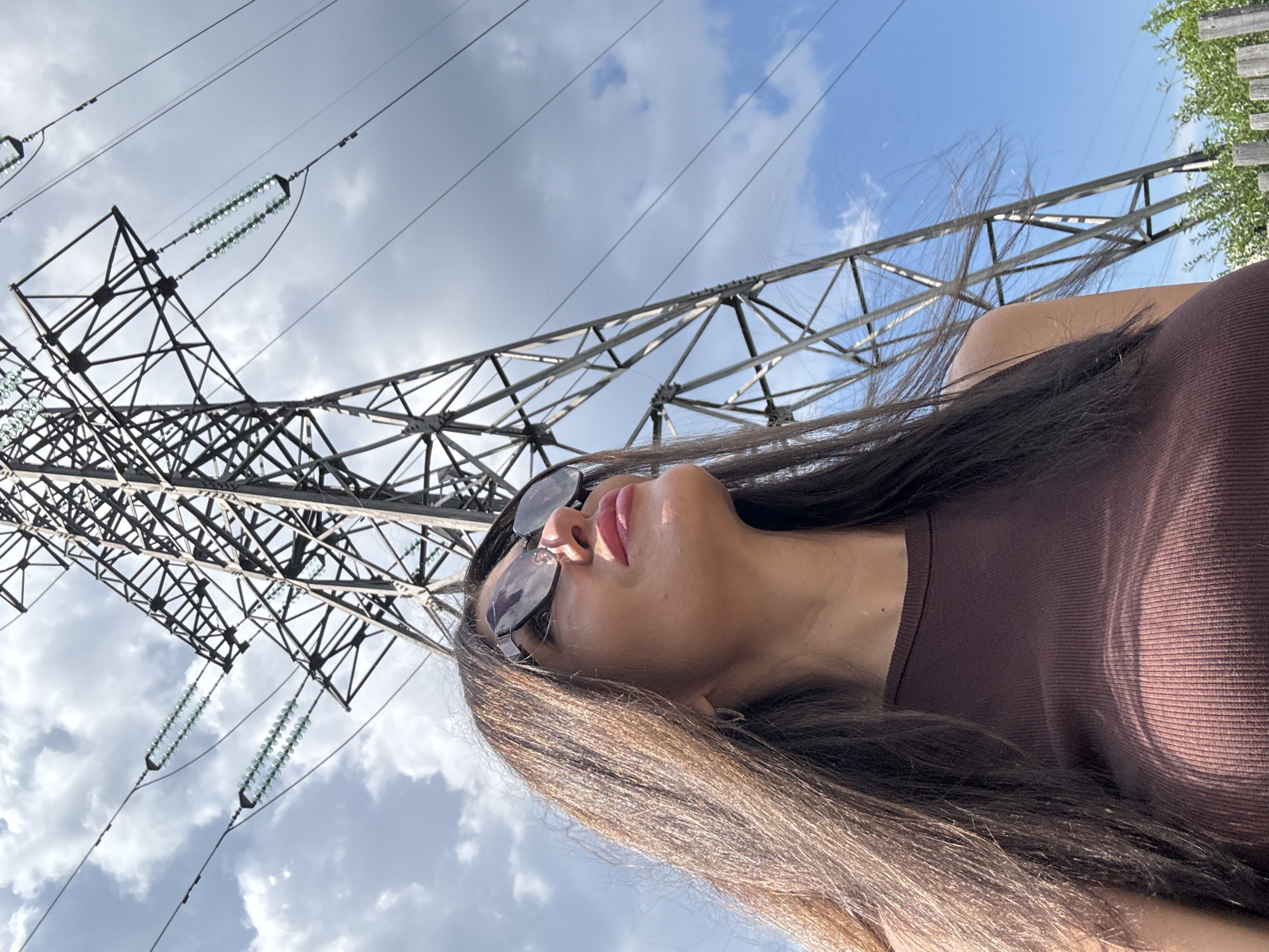 A low-angle medium shot shows a woman with long dark hair wearing sunglasses and a brown ribbed tank top, looking up towards a towering electrical pylon. The pylon, a lattice structure of steel, dominates the upper portion of the frame, with multiple power lines extending from it against a sky filled with scattered clouds. The woman's face is partially visible from the side, her lips slightly parted, and she appears to be outdoors. In the background, a line of trees and a wooden fence can be seen, suggesting a rural or semi-rural setting. The image does not contain any specific landmarks to identify its exact location in Chaikovsky, Russia.