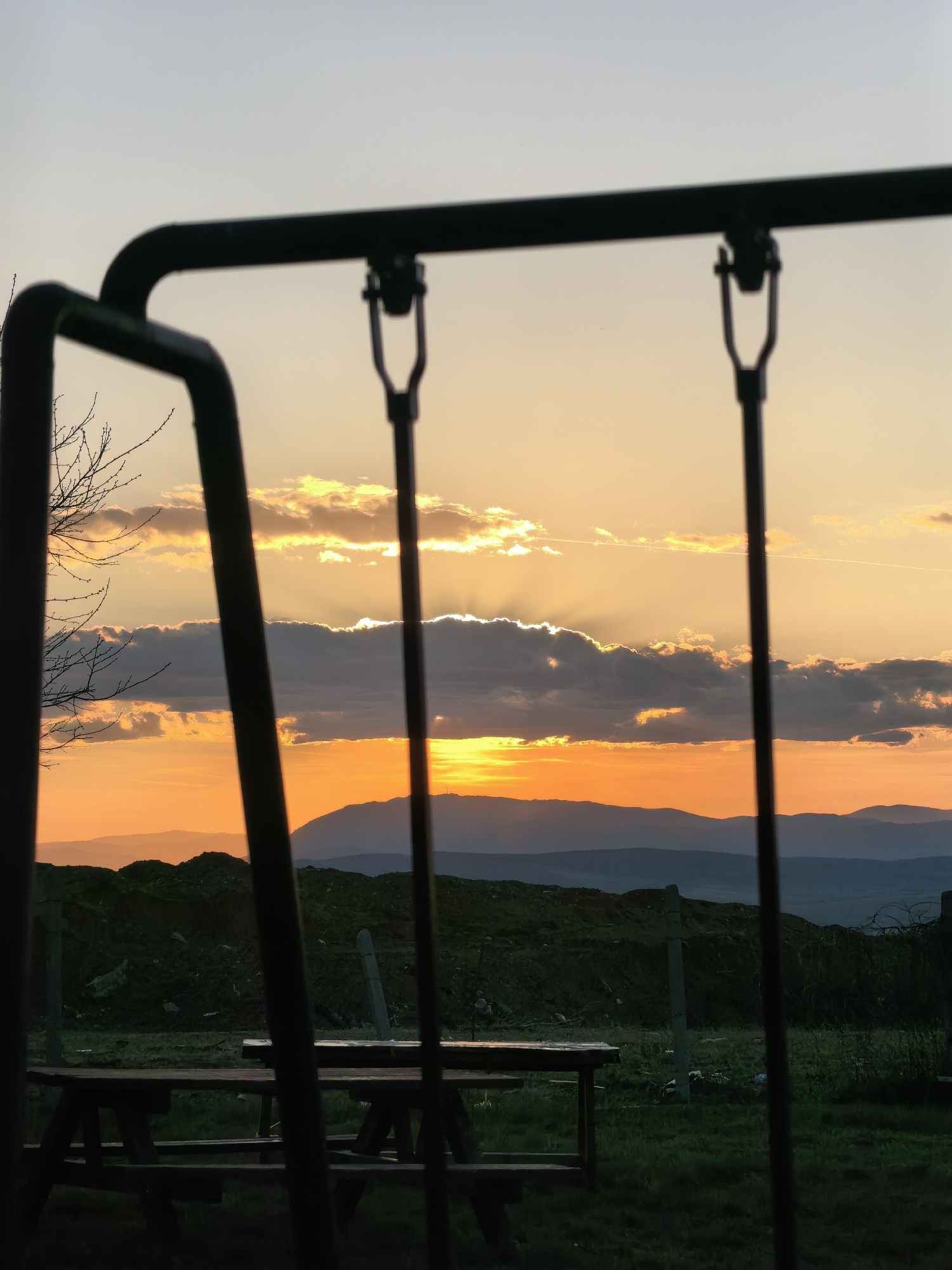 A low-angle shot of a park with a metal swing set partially in the foreground. Behind the swing set, there is a picnic table on a grassy area. The landscape features low hills and mountains in the distance. The sky is a sunset with orange and yellow hues and some clouds. The city is Unknown, Unknown.