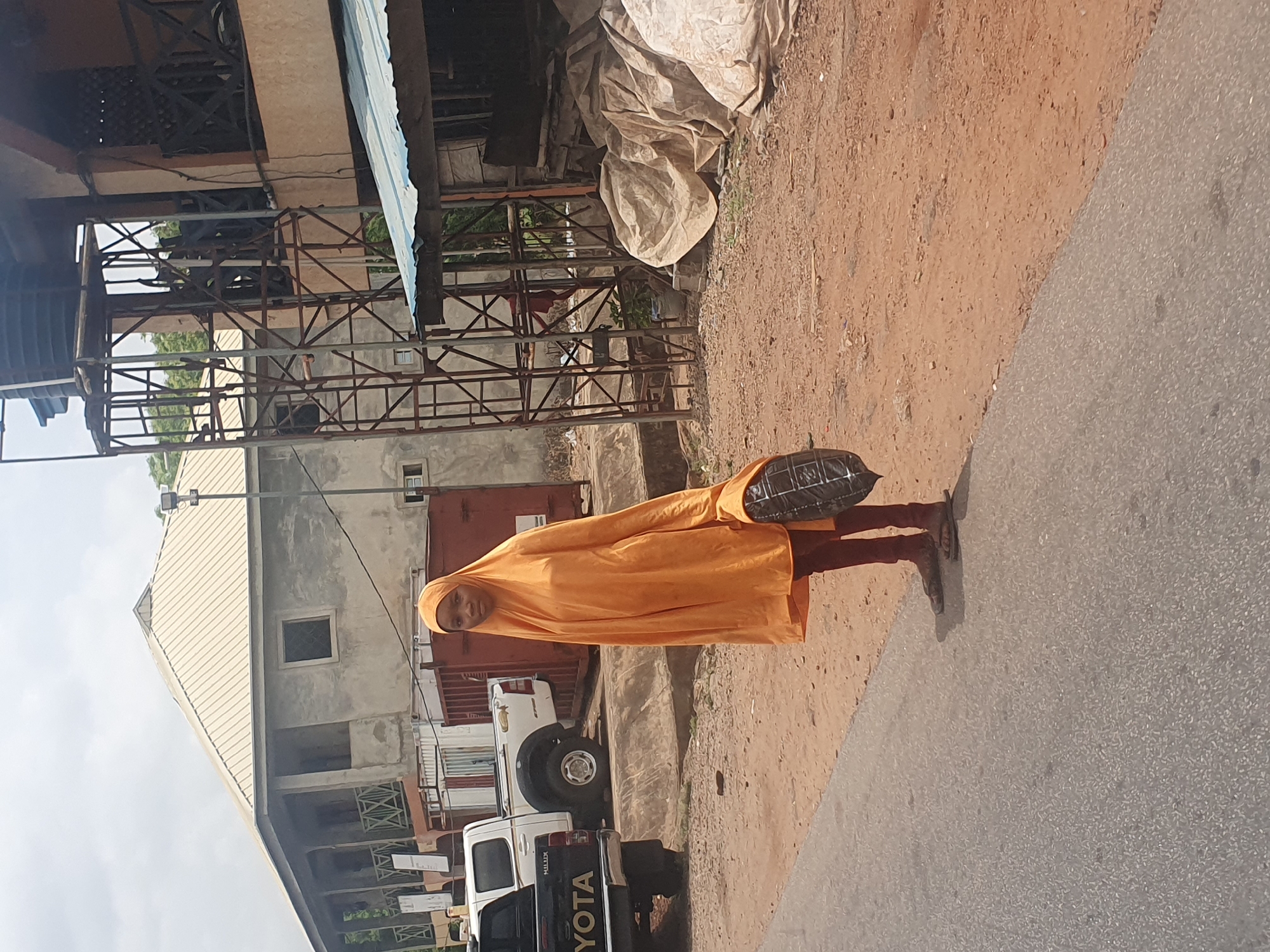 A woman wearing an orange hijab and dress walks on the side of a street in Lokoja, Nigeria. She carries a black bag. In the background, there is a building with a metal frame structure, a beige-colored building, a white Toyota Hilux pickup truck, and tarpaulin-covered items. The street surface is a mix of red dirt and asphalt.