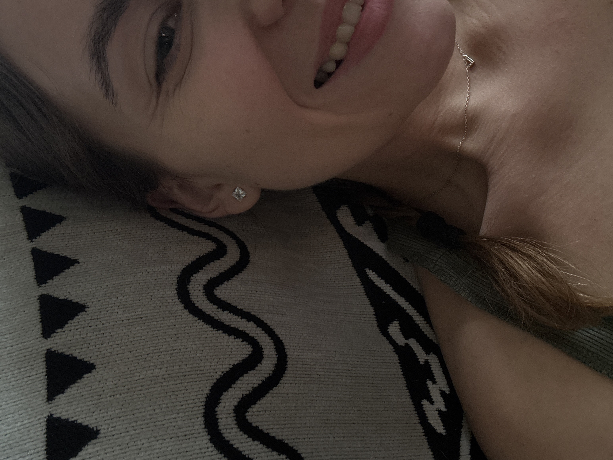 The image shows a close-up of a woman's face and upper body, with her head turned towards the camera and her body partially obscured by a textured blanket with a black geometric pattern. She has light brown hair styled in a braid, a silver stud earring in her left ear, and is wearing a delicate gold necklace with a small heart pendant. Her right eye is visible, and she is smiling, revealing her teeth. The background is a textured blanket with a repeating pattern of black triangles and wavy lines. The lighting suggests an indoor setting. There is no identifiable information or context provided in the image to place it within the city of Zheleznogorsk, Russia.
